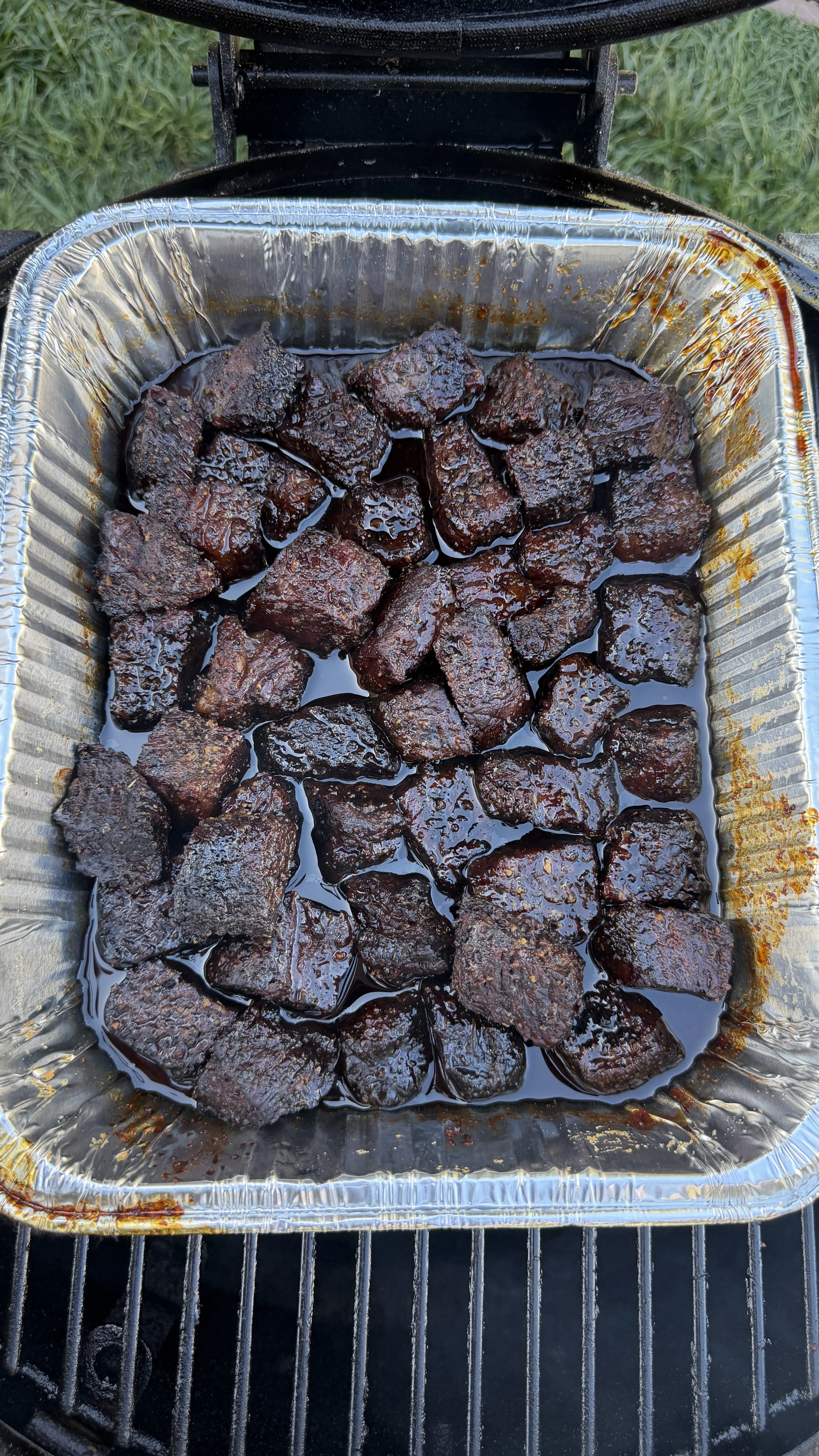 Dark, caramelized smoked molasses coffee beef rib burnt ends in aluminum foil pan on outdoor grill