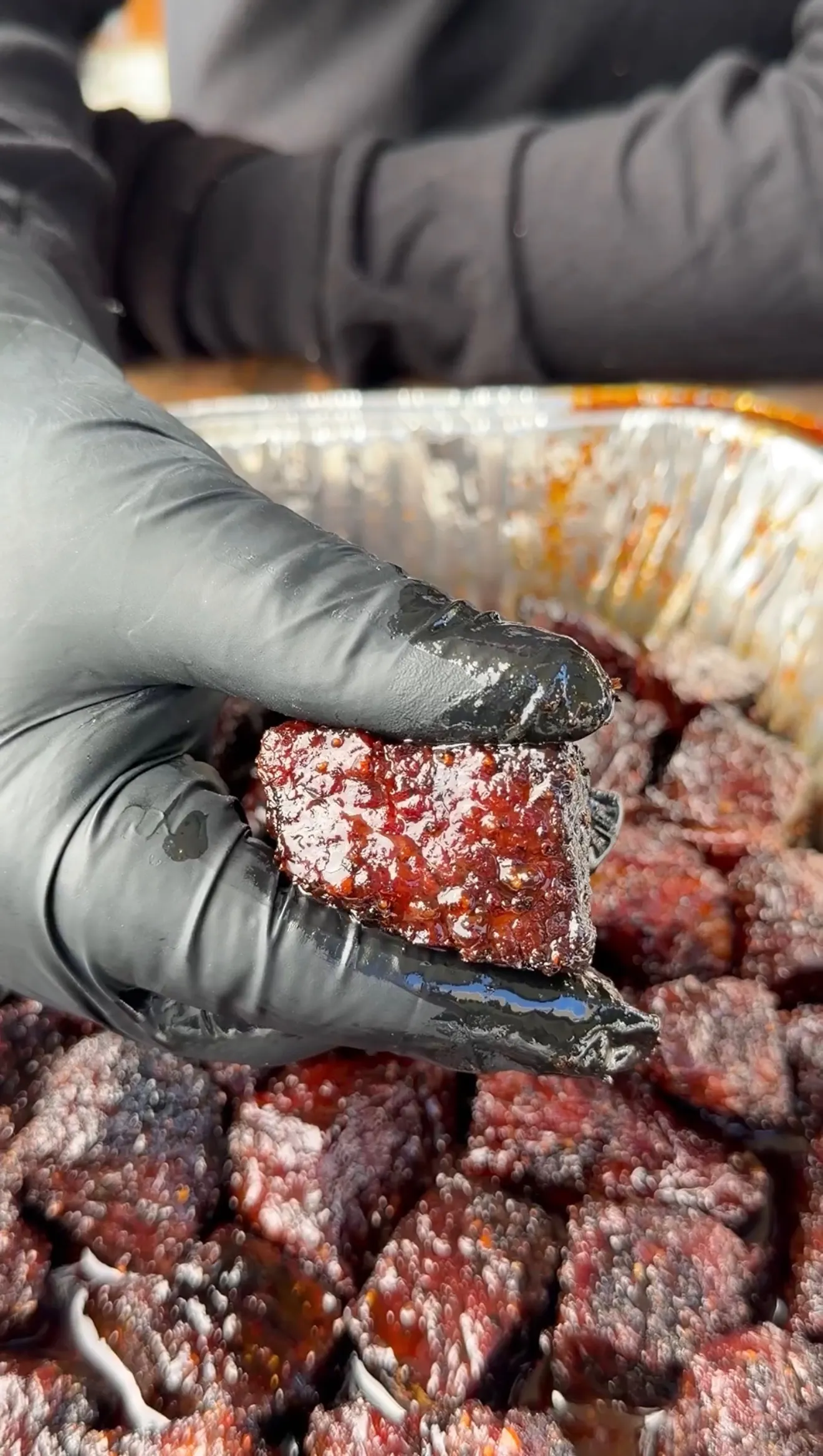 Hand in black glove picking up a caramelized smoked beef rib burnt end from a metal bowl filled with cubed meat