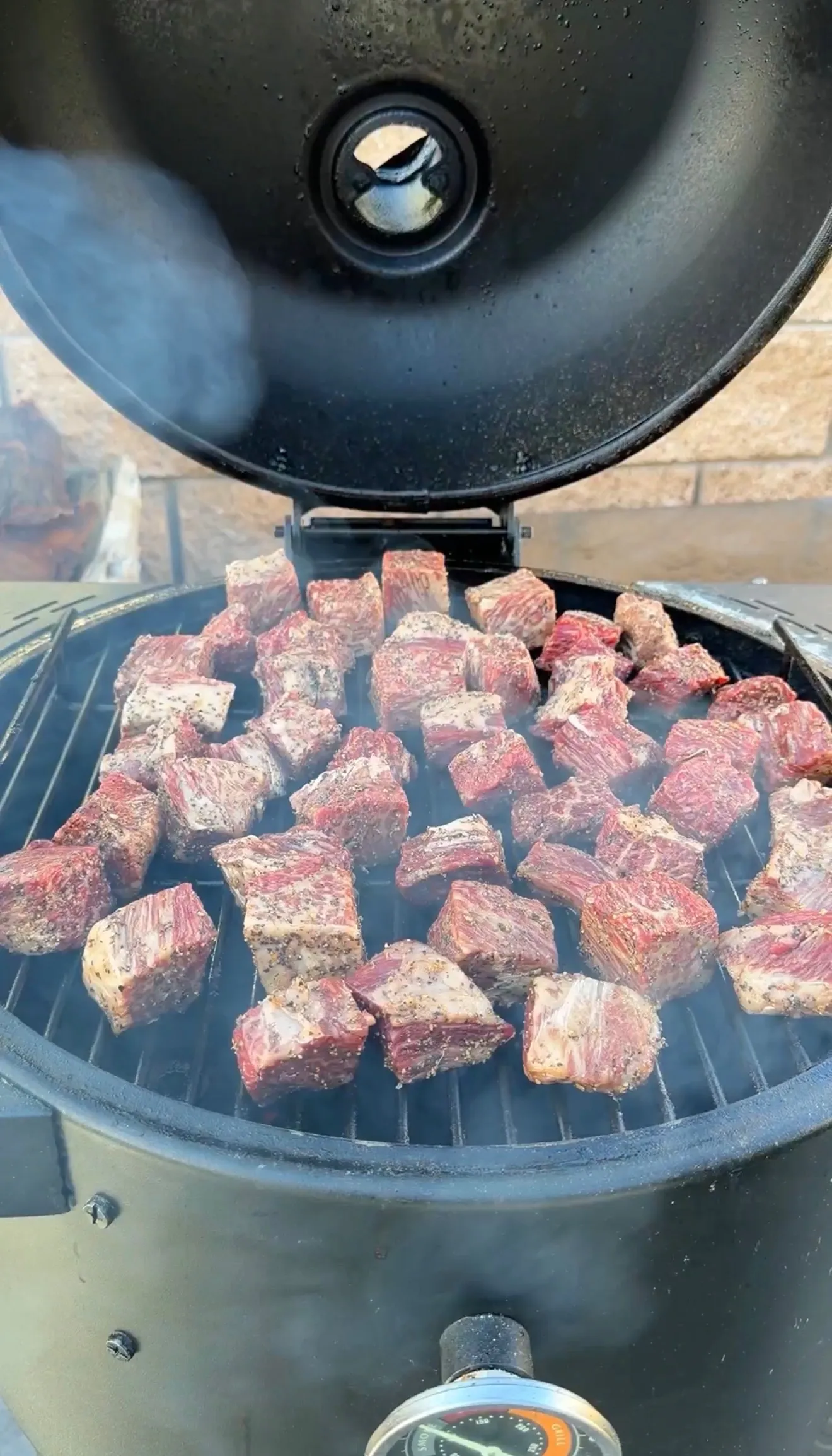 Cubed smoked molasses coffee beef rib burnt ends cooking on a round grill grate with lid open