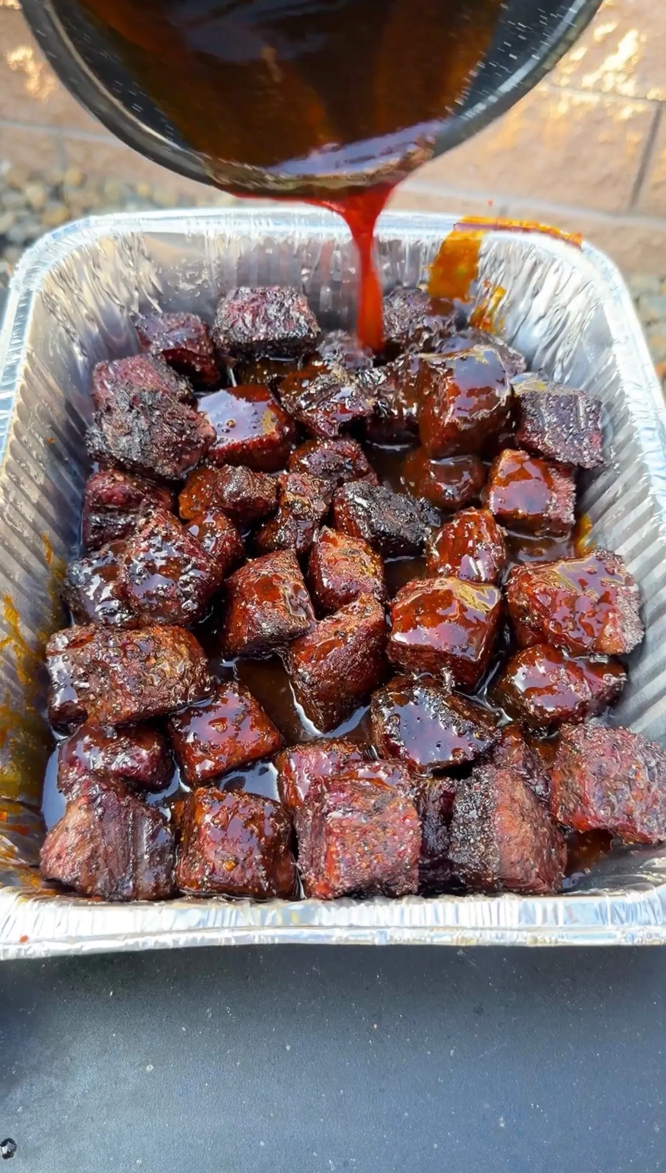 Glazed smoked beef rib burnt ends in aluminum tray being drizzled with molasses coffee barbecue sauce