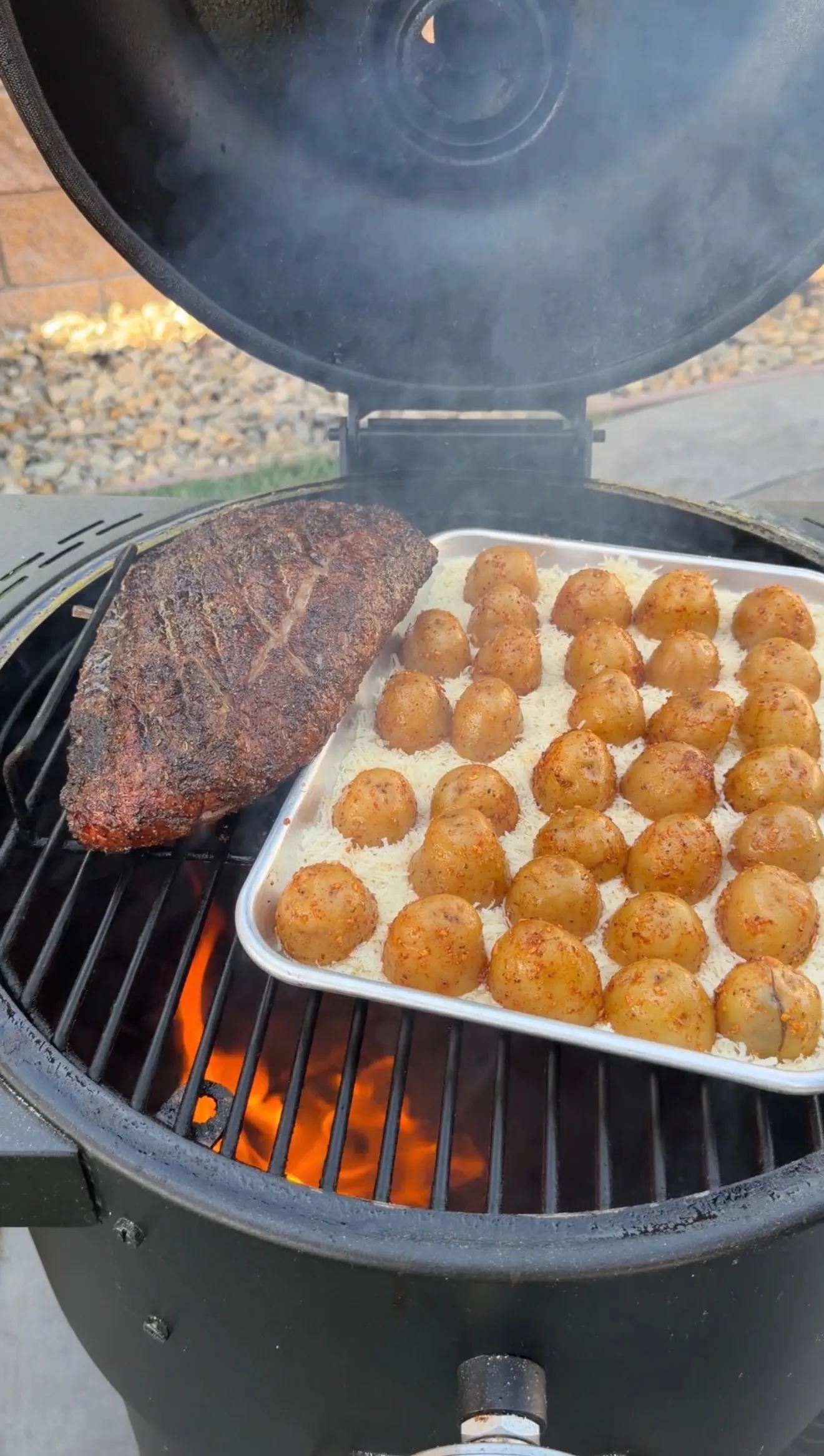 Smoked picanha roast and crispy parmesan potatoes cooking on a charcoal grill with visible flames and smoke