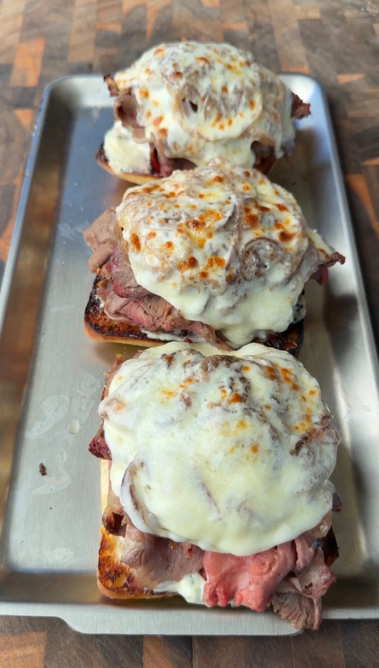 Three garlic herb butter smoked prime rib French dip sandwiches topped with melted cheese on a metal tray