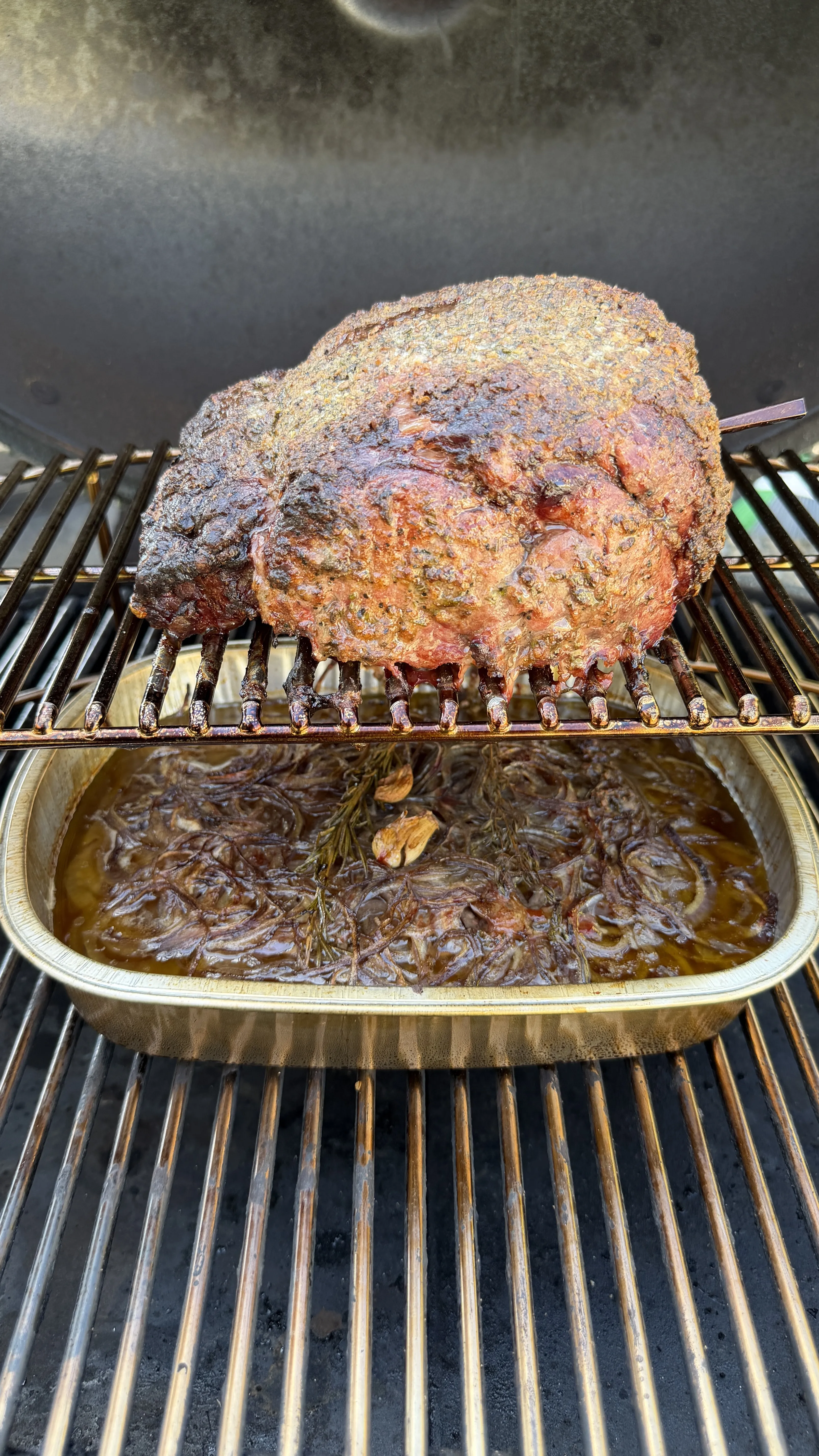 Herb-crusted smoked prime rib roast on grill grates with au jus dripping pan underneath