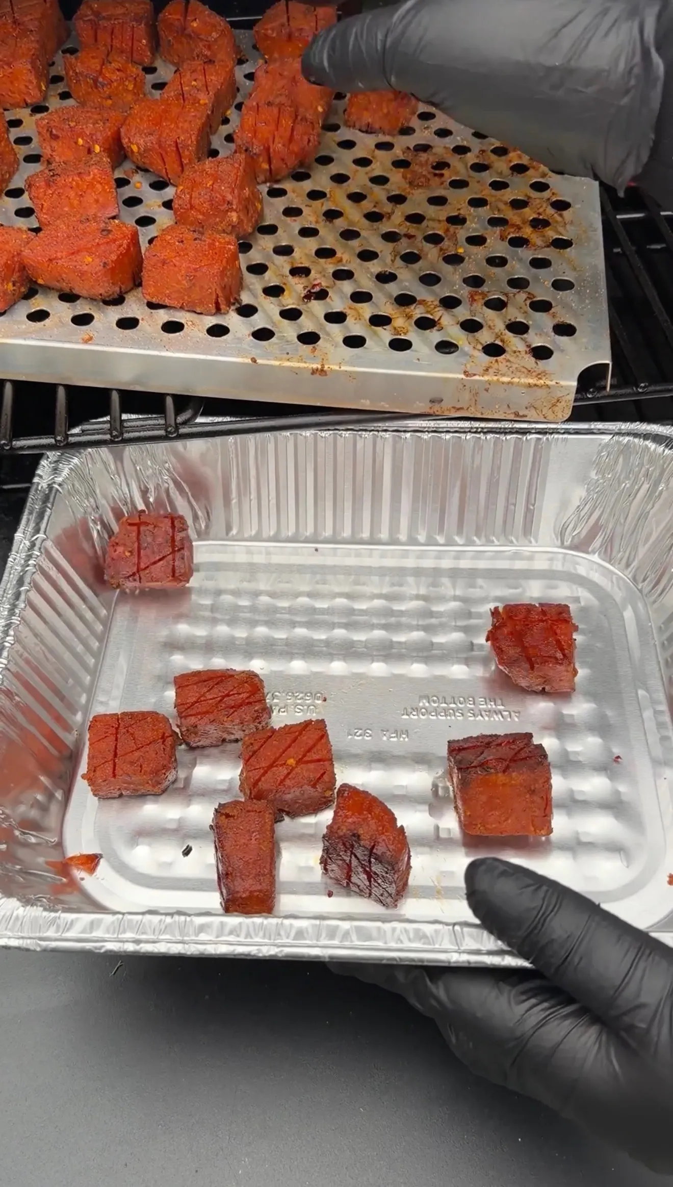 Caramelized spam burnt ends being transferred from a perforated smoker tray into a disposable aluminum pan