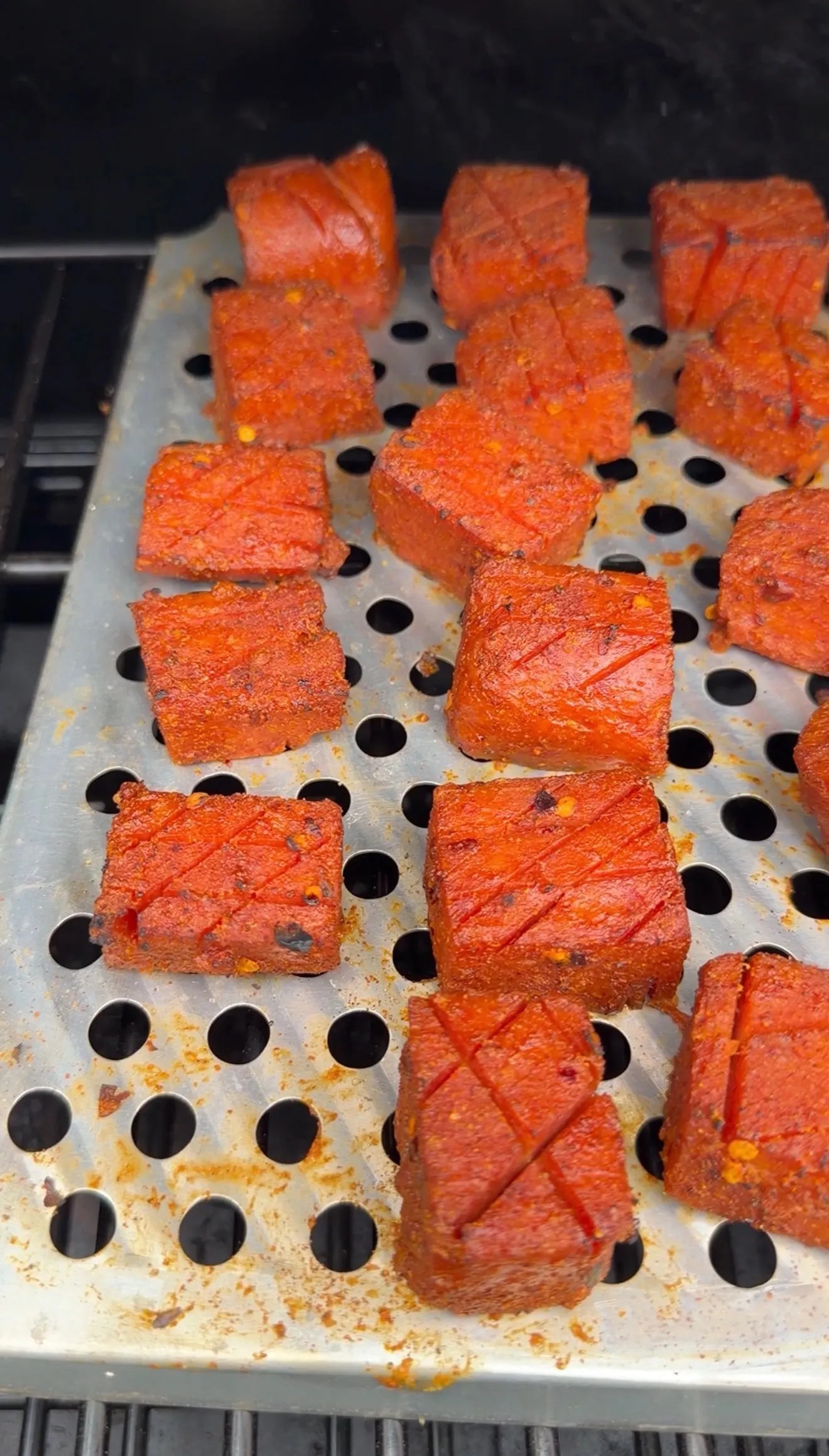 Seasoned Spam burnt ends with crosshatch scoring, coated in red spice rub, arranged on a perforated smoker tray