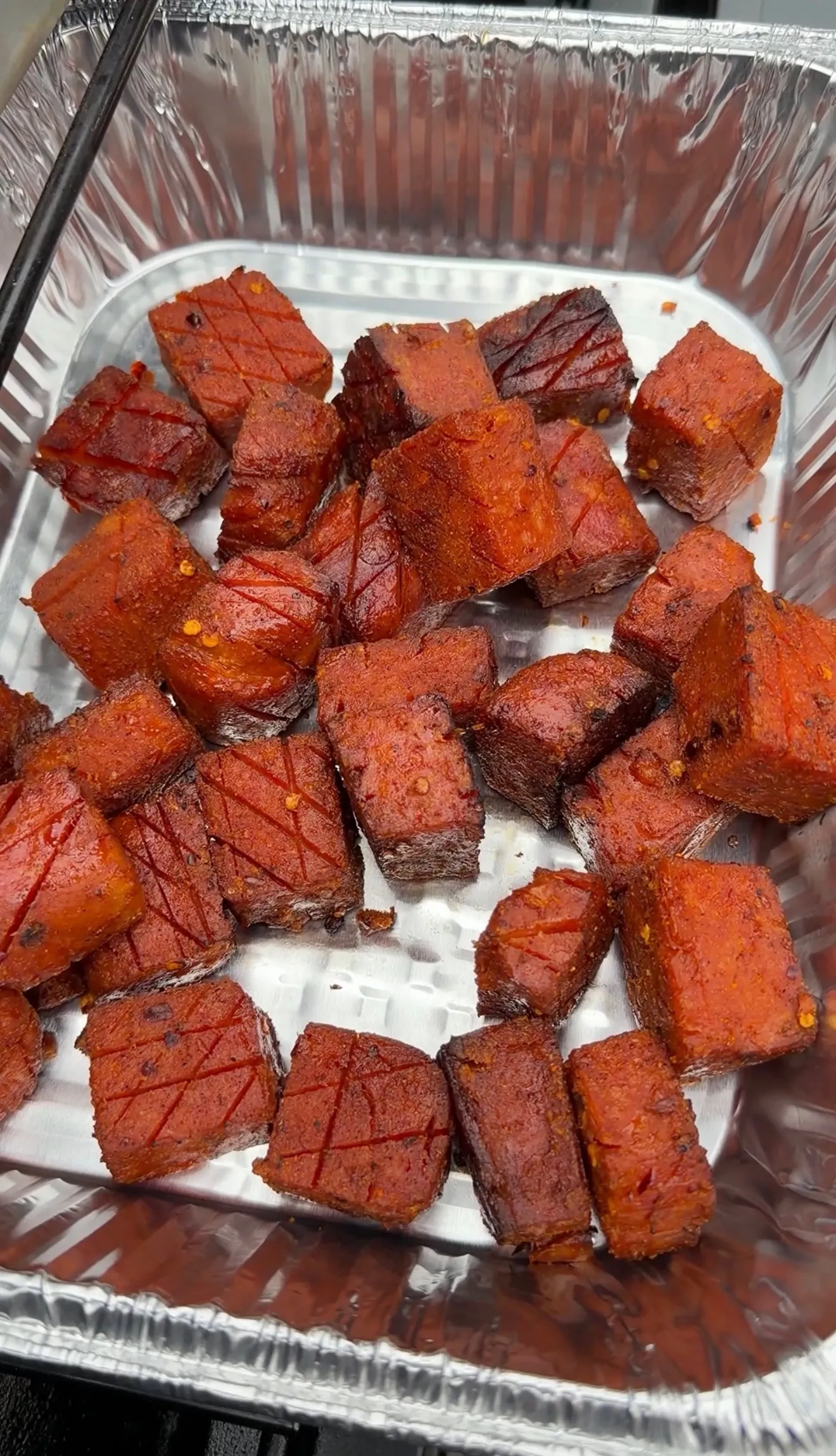 Caramelized spam burnt ends cubes with spicy red glaze served in a disposable aluminum foil pan