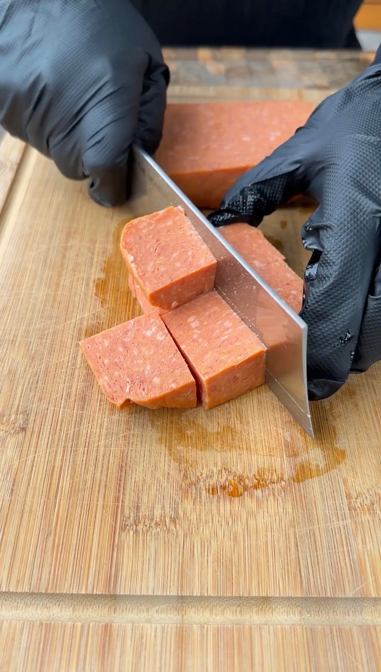 Gloved hands cutting a block of Spam into cubes on a wooden cutting board for burnt ends recipe