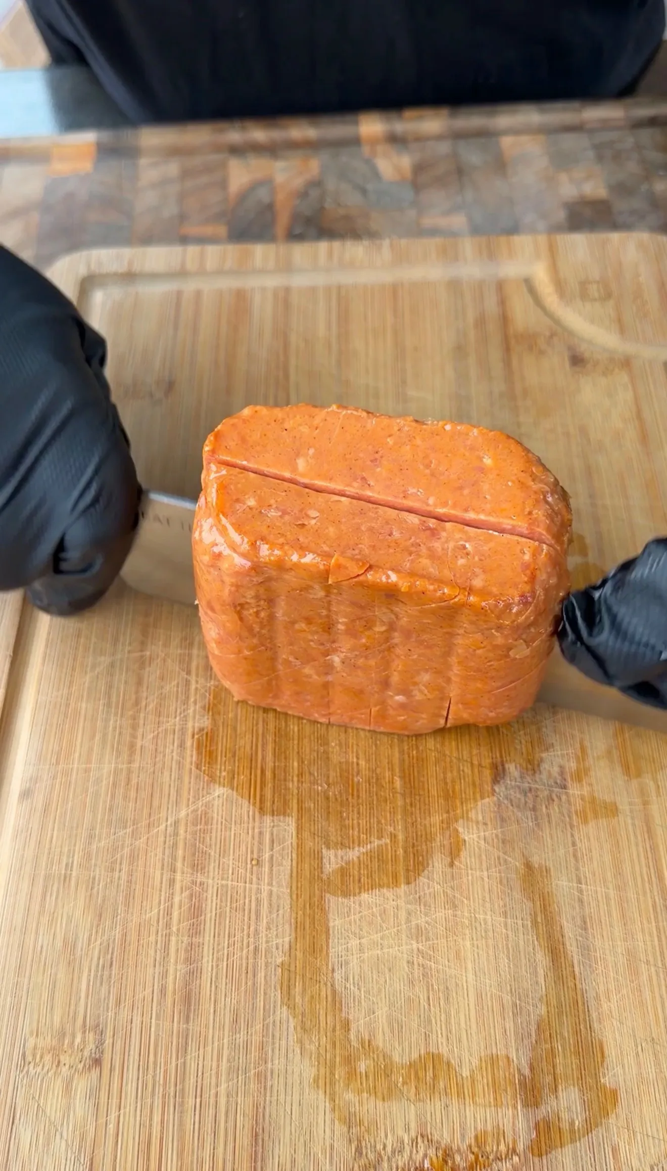 Hands in black gloves scoring a block of seasoned Spam with a knife on a wooden cutting board