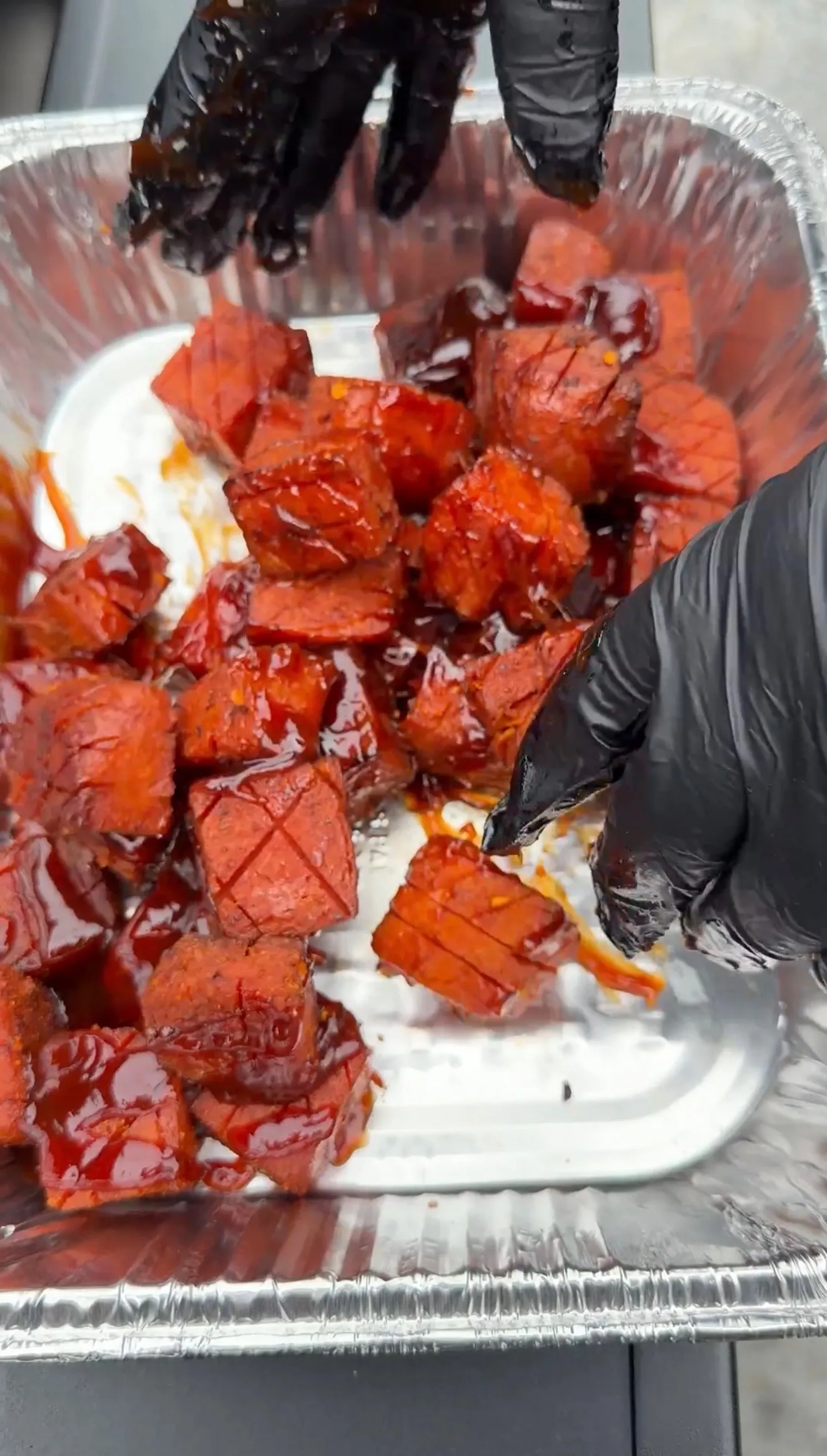 Caramelized spam burnt ends coated in sticky BBQ sauce in an aluminum pan, tossed with black gloved hands