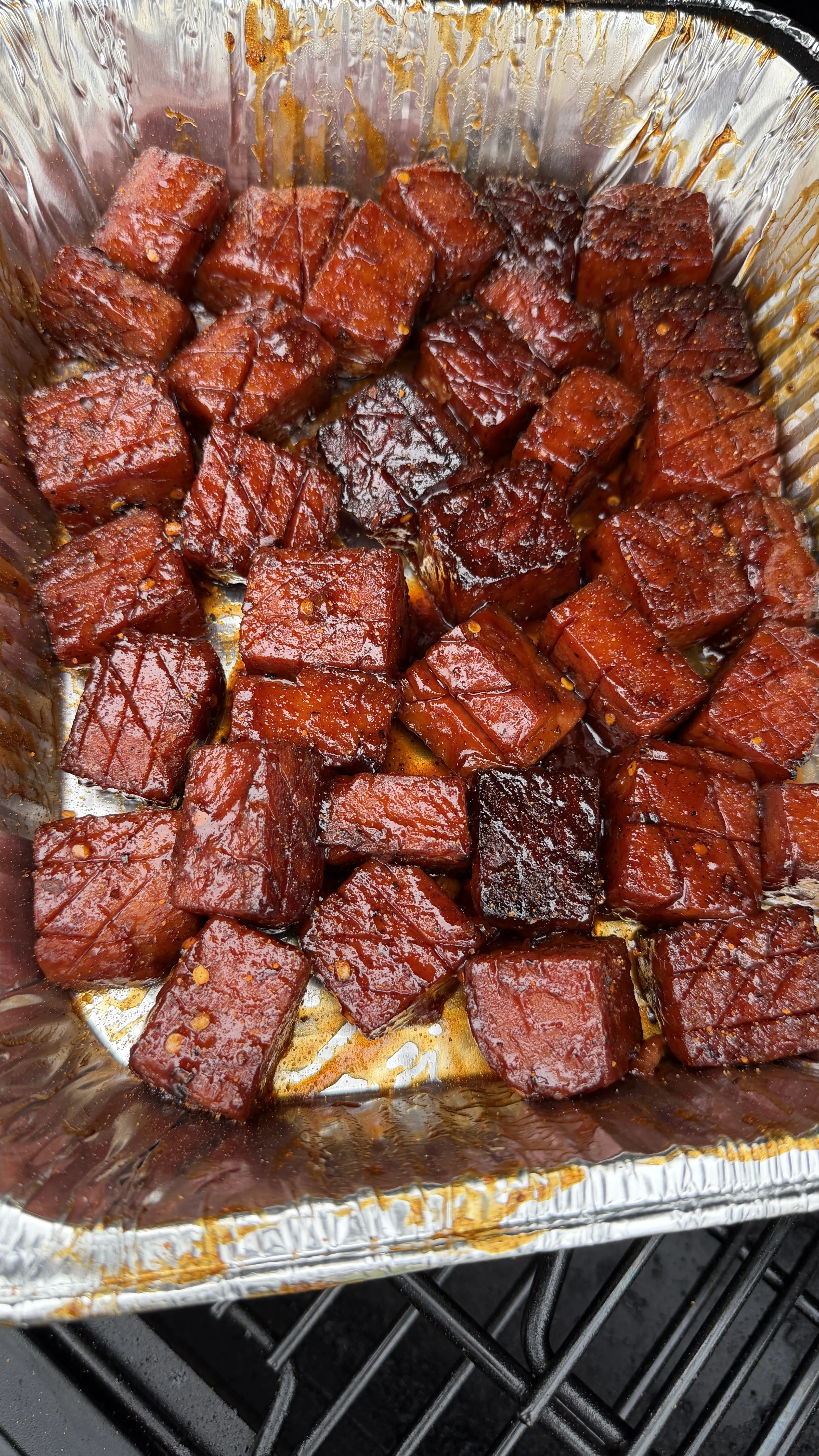 Caramelized spam burnt ends coated in sticky BBQ glaze in an aluminum foil pan on a smoker grill