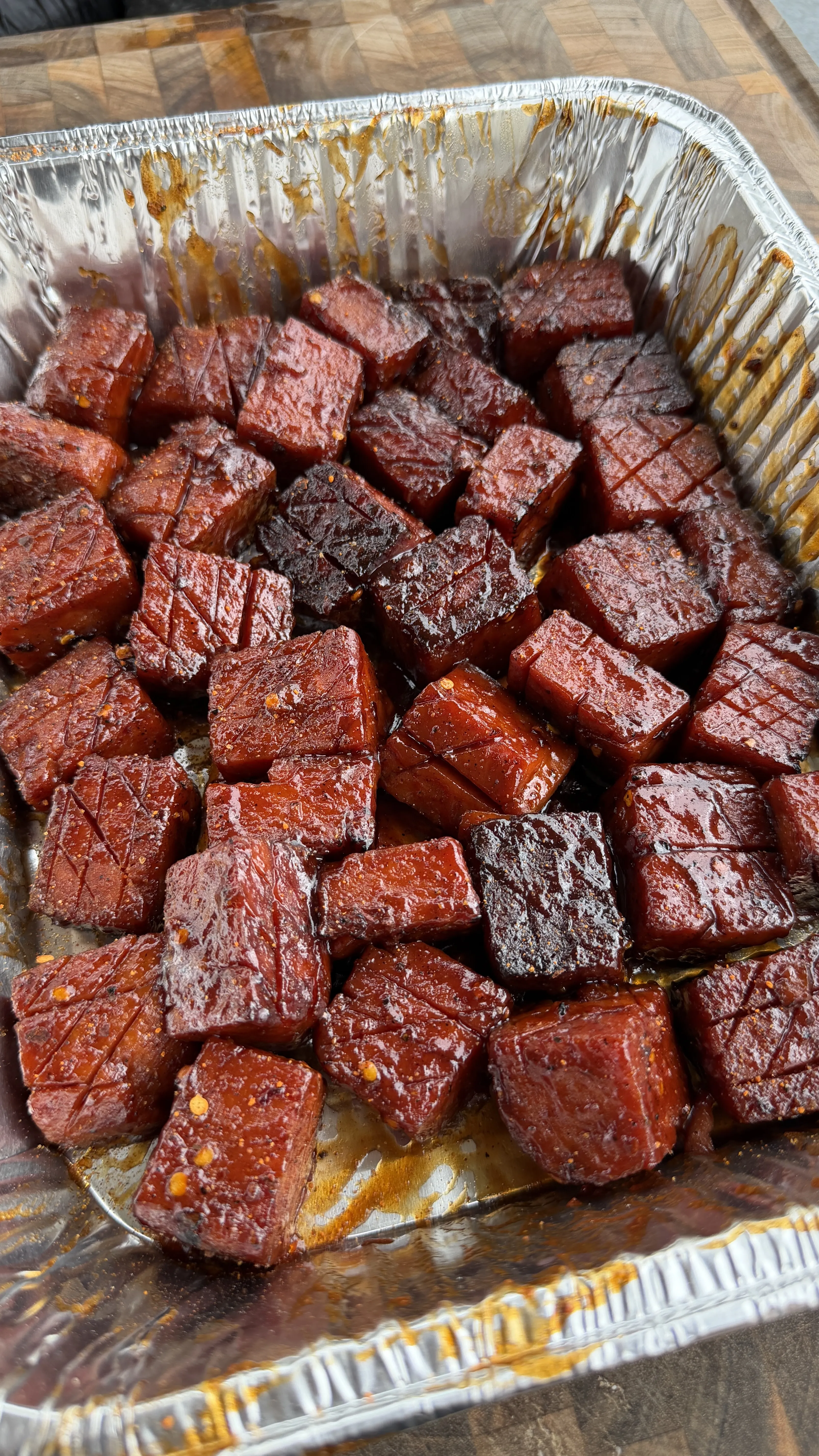 Caramelized spam burnt ends coated in sticky BBQ glaze arranged in a disposable aluminum foil pan