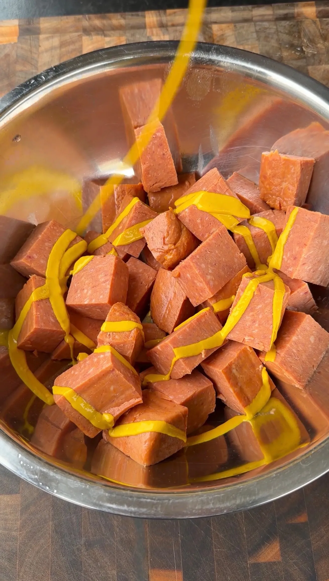 Cubed Spam pieces coated with yellow mustard in a stainless steel mixing bowl, ready for burnt ends preparation