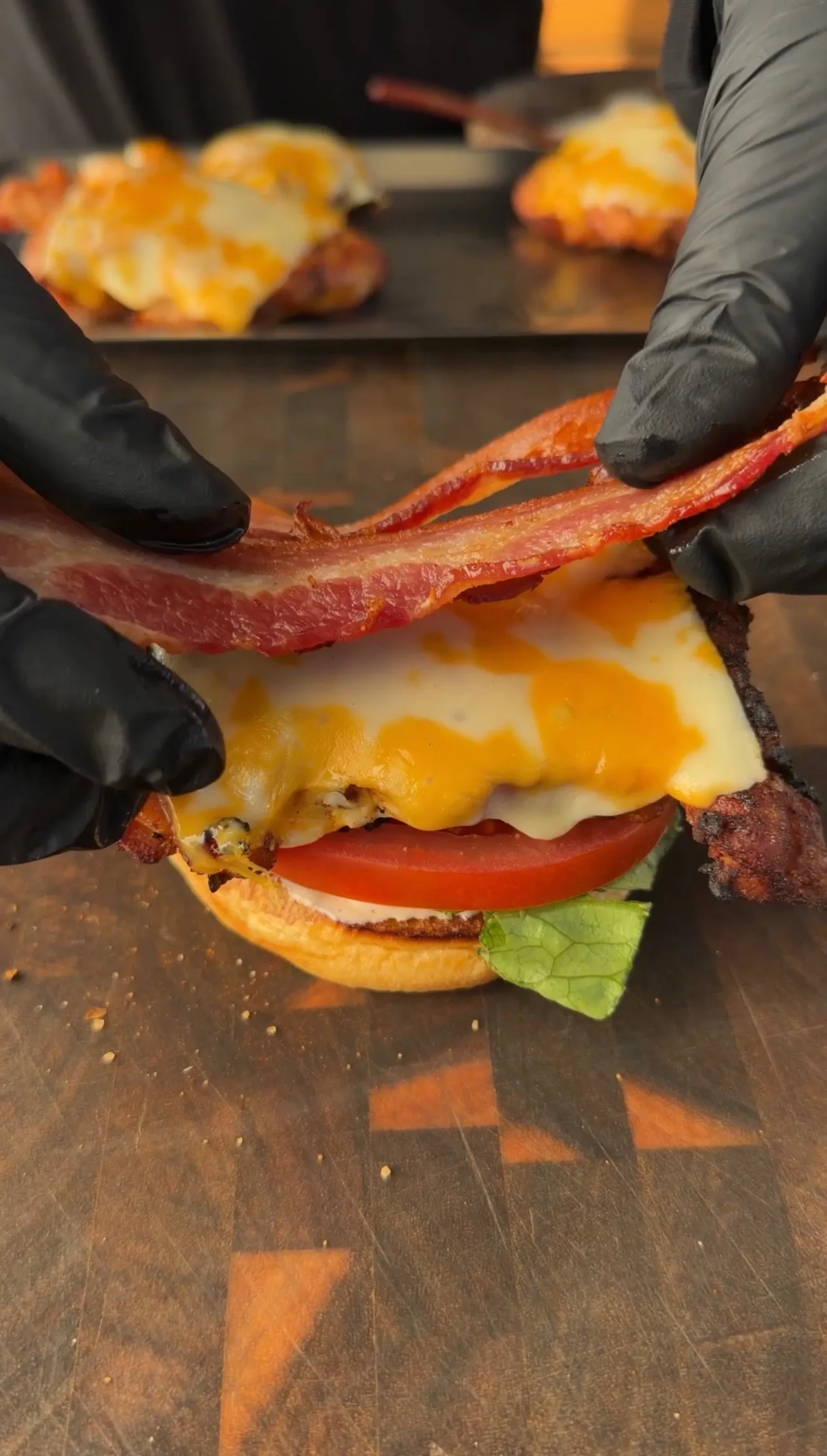 Hands placing crispy bacon on a spicy chicken BLT sandwich with melted cheese, tomato, and lettuce on a wooden board