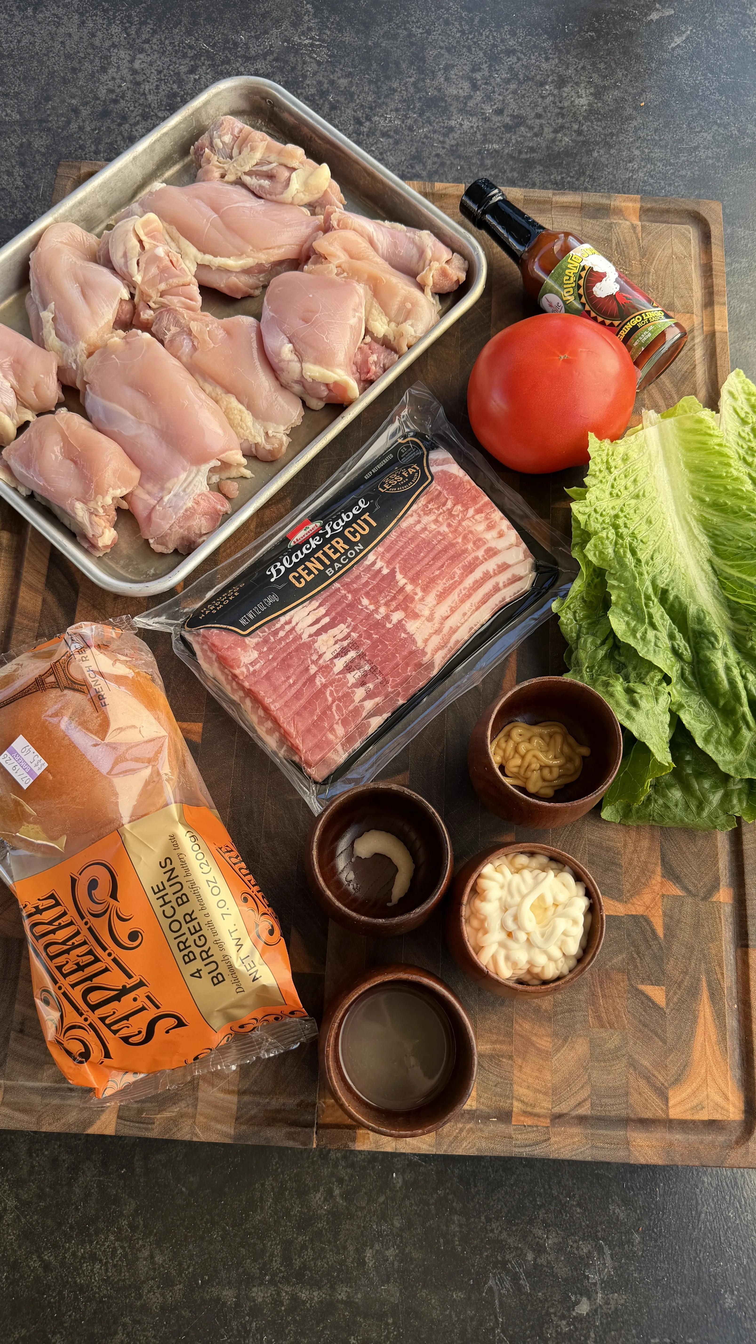 Raw chicken thighs, bacon, brioche buns, tomato, lettuce, hot sauce, mustard, and mayo on a wooden board
