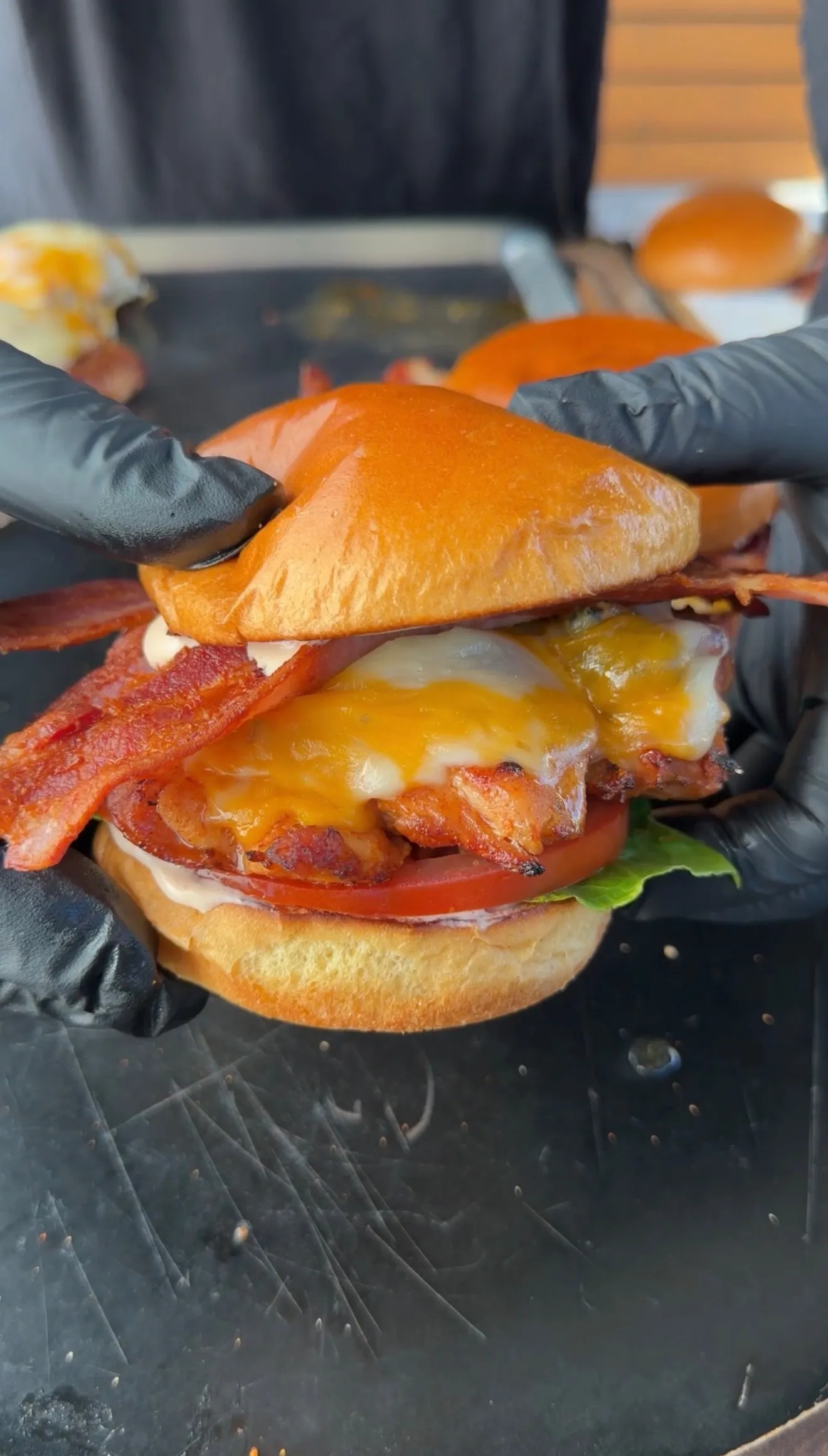 Hands holding a spicy chicken BLT sandwich with melted cheese, crispy bacon, and tomato on a brioche bun