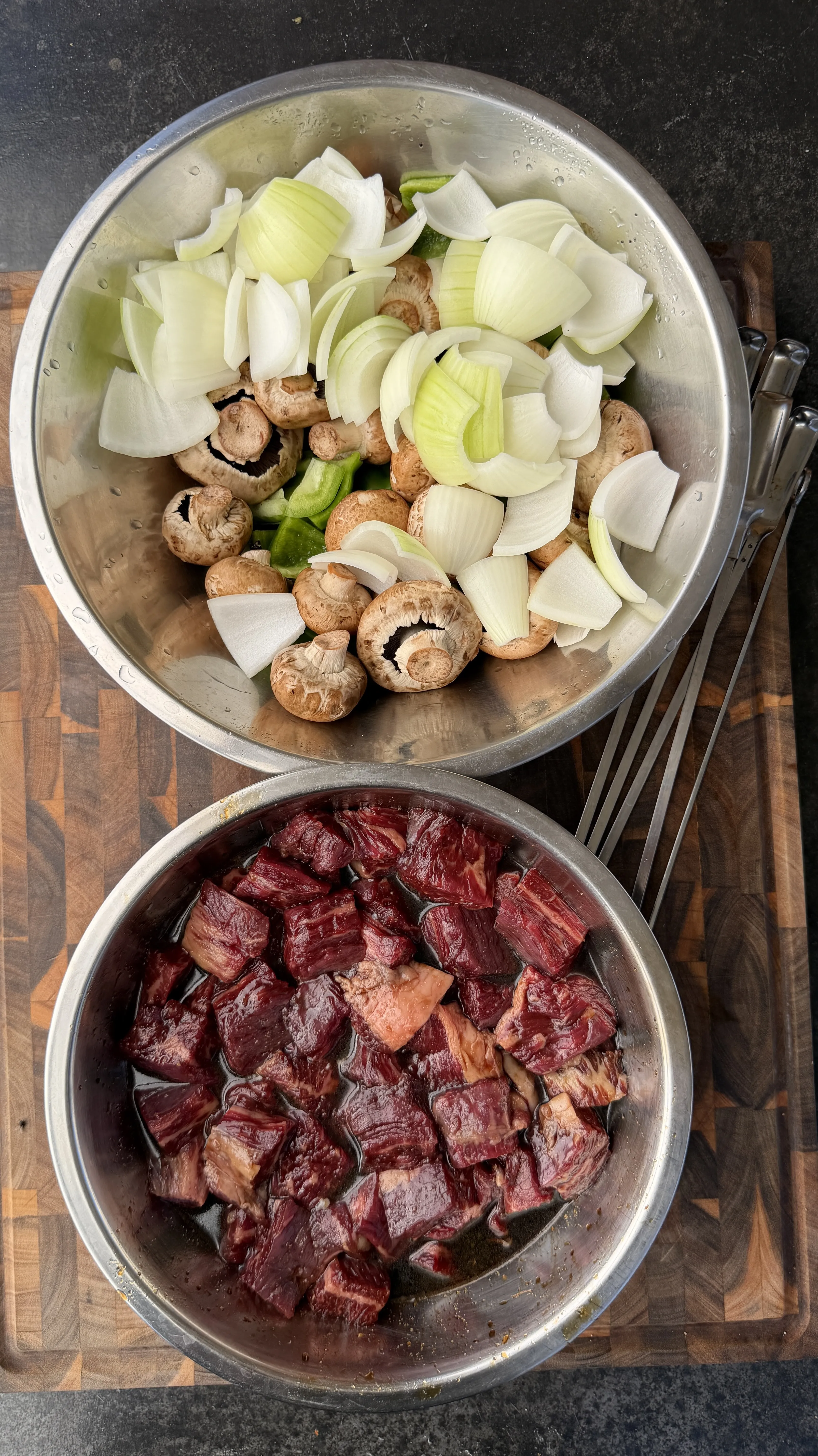 Raw cubed beef and sliced vegetables in metal bowls on wooden cutting board for teriyaki beef skewers