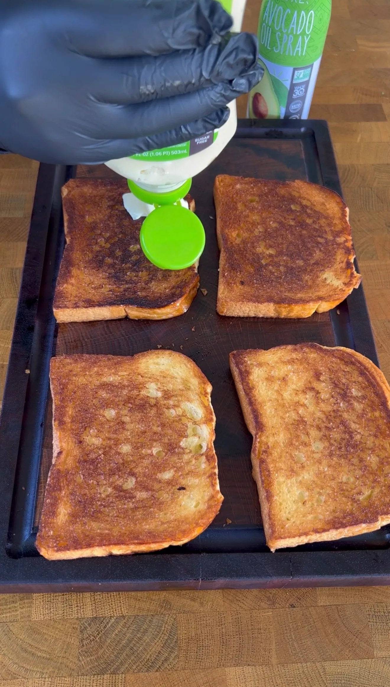 Four golden-brown toasted bread slices on black griddle with hand spraying avocado oil for BLT preparation
