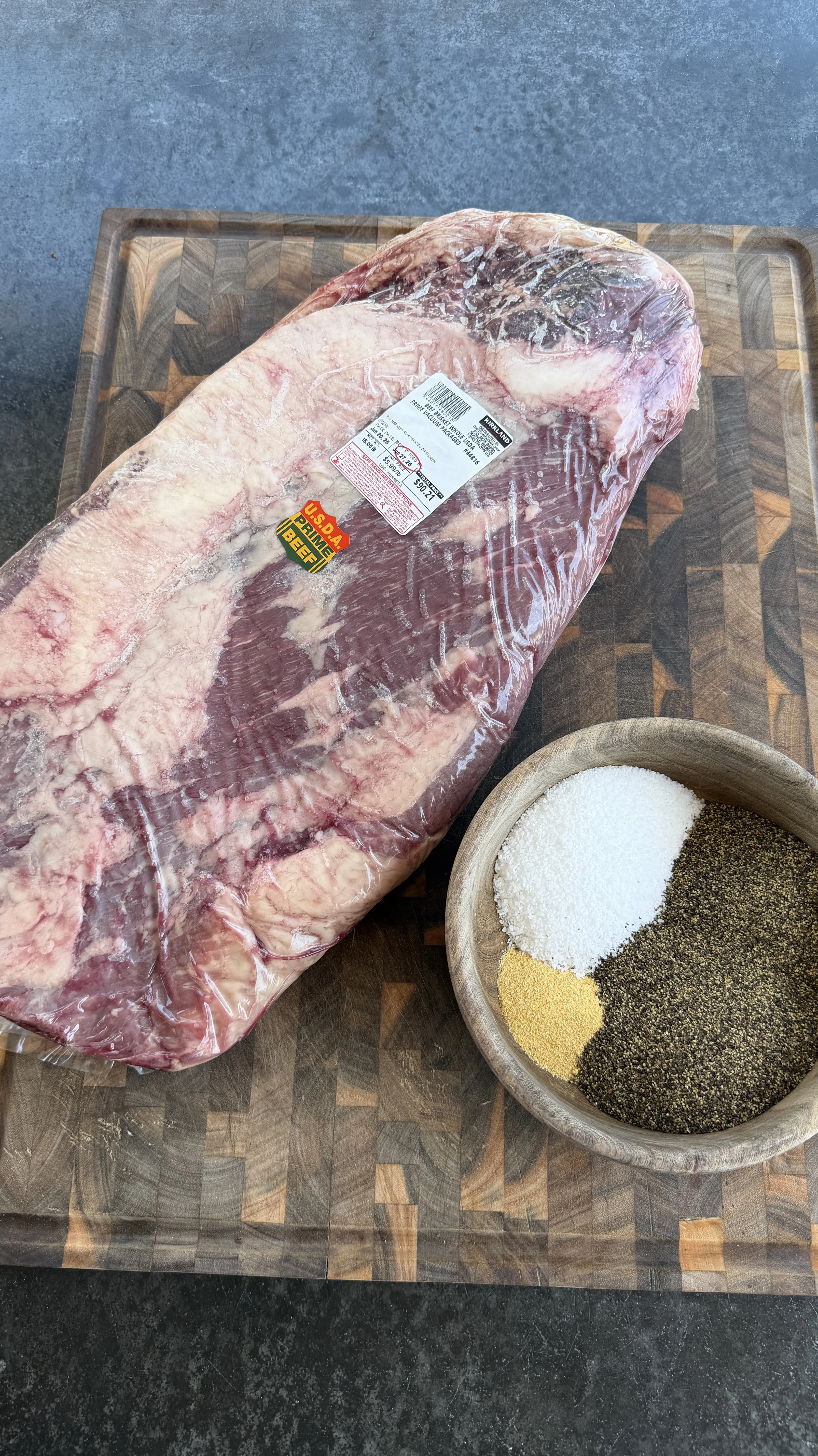 Whole USDA Prime beef brisket in packaging on cutting board with bowl of salt, pepper, and garlic seasoning rub