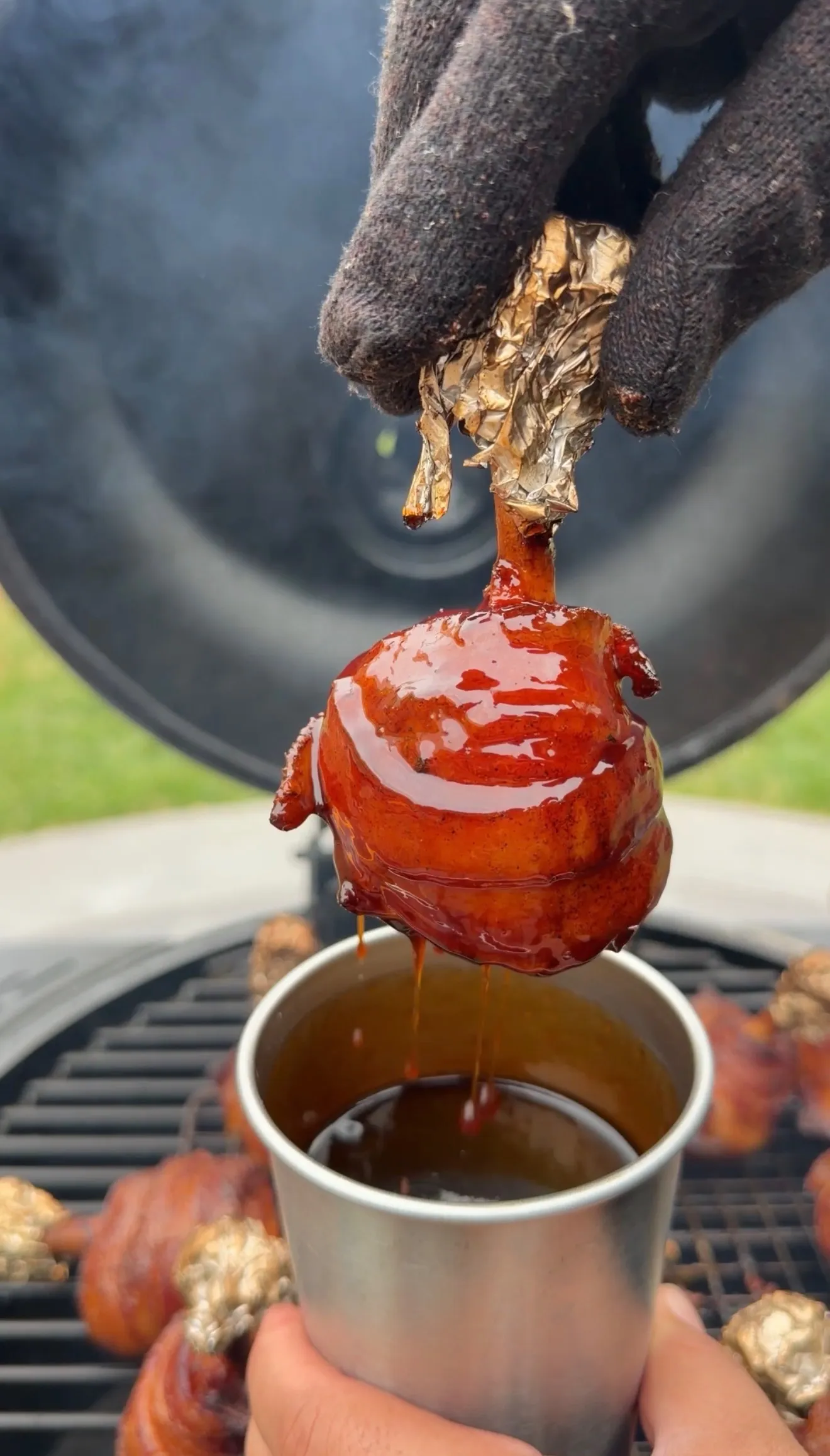 Glazed bacon wrapped chicken lollipop being dipped in BBQ sauce over a metal cup on a smoker grill