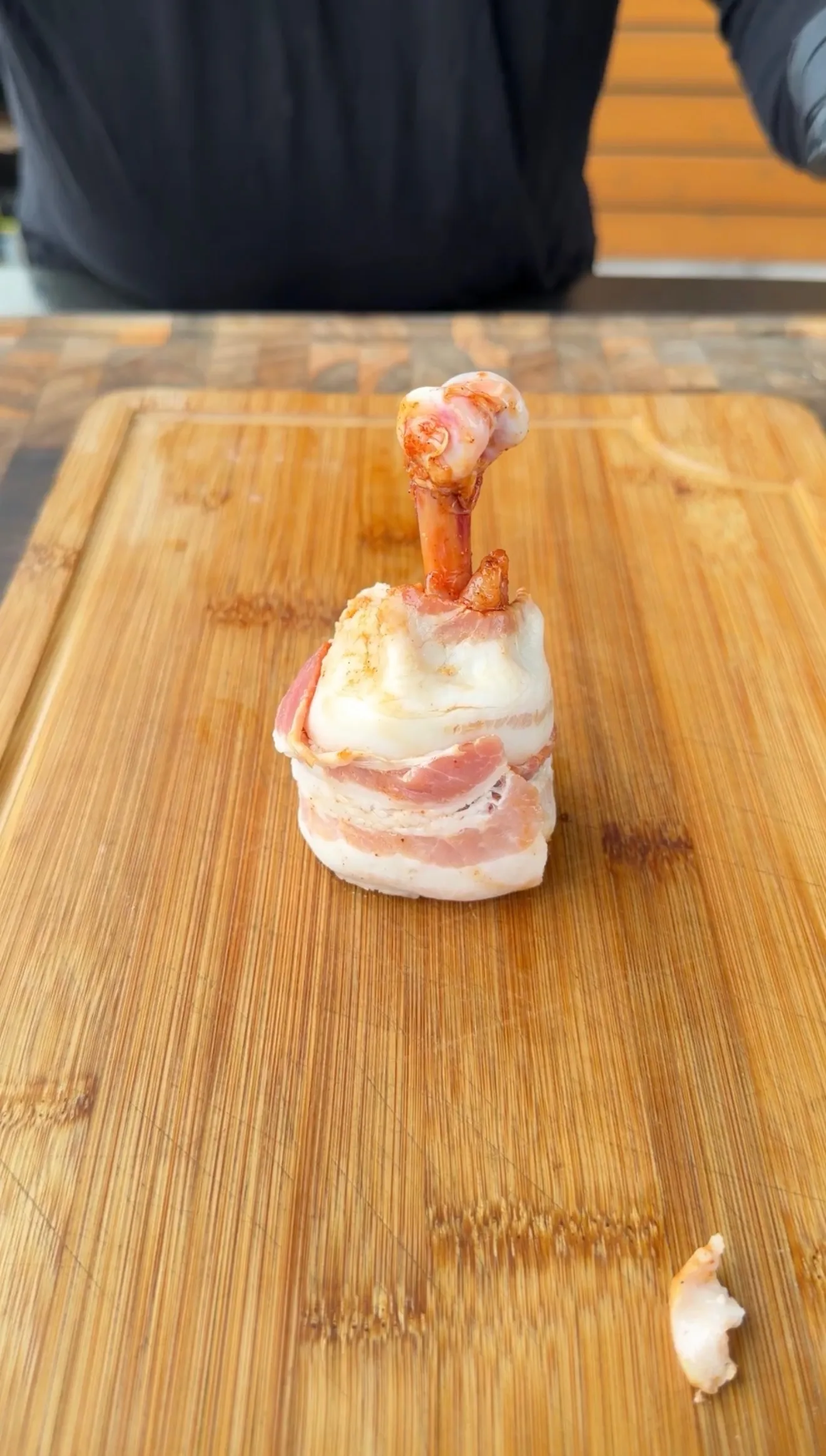 Raw bacon-wrapped chicken lollipop standing upright on a wooden cutting board, ready to cook