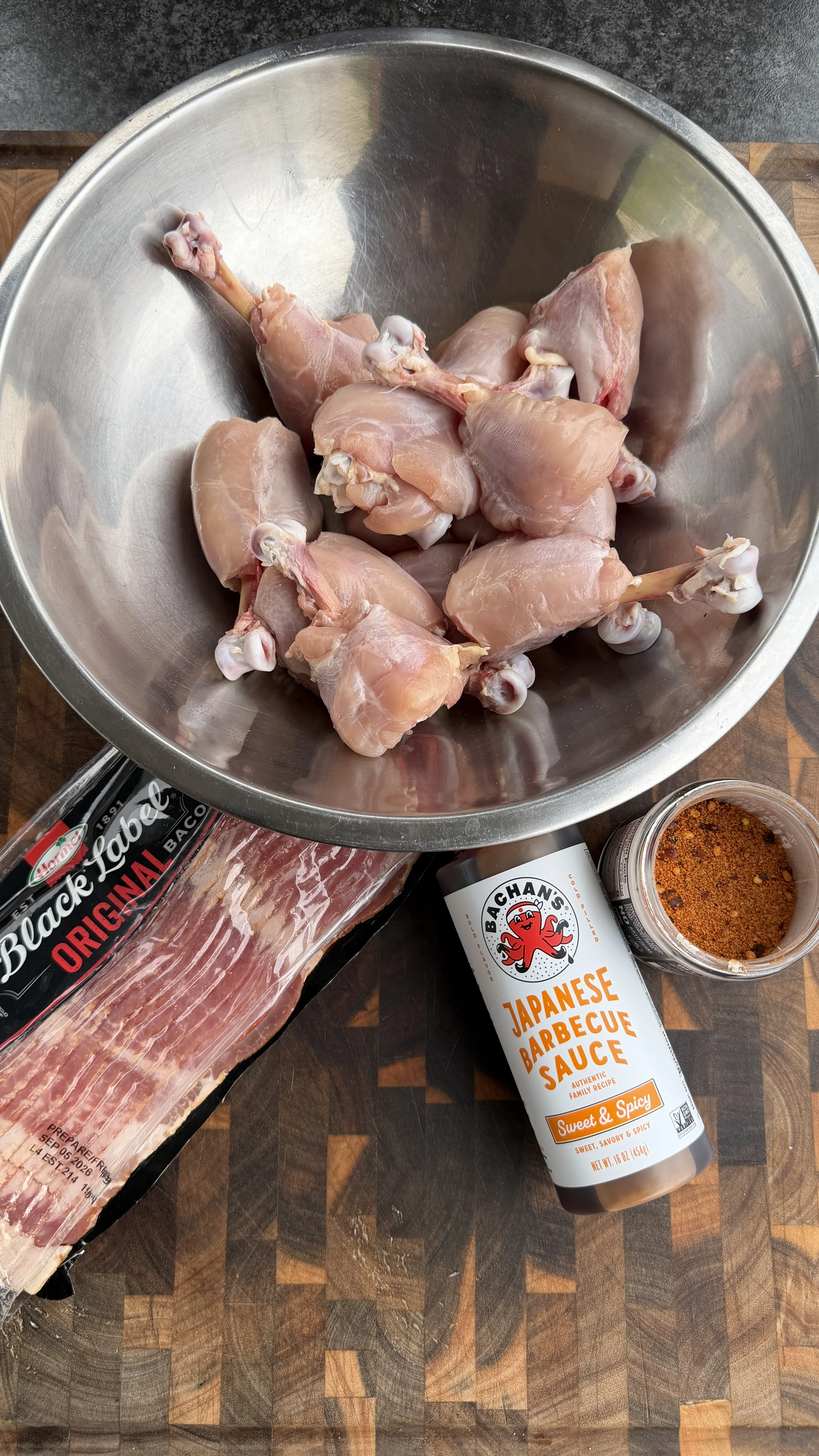 Raw chicken lollipops in a steel bowl with bacon, Bachan's Japanese BBQ sauce, and spice rub on a wood board