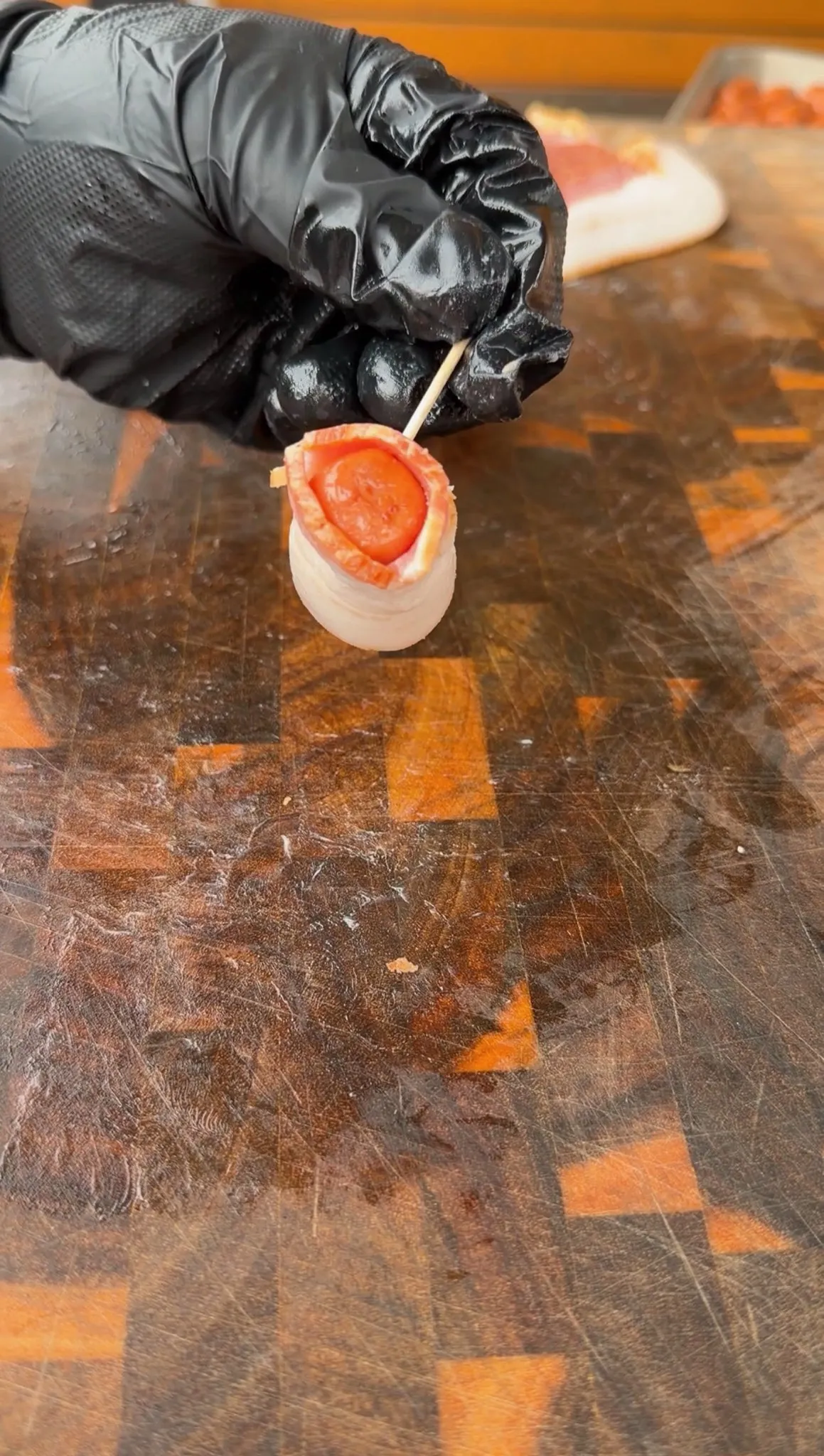 Gloved hand holding a bacon-wrapped little smokie secured with a toothpick over a wooden cutting board