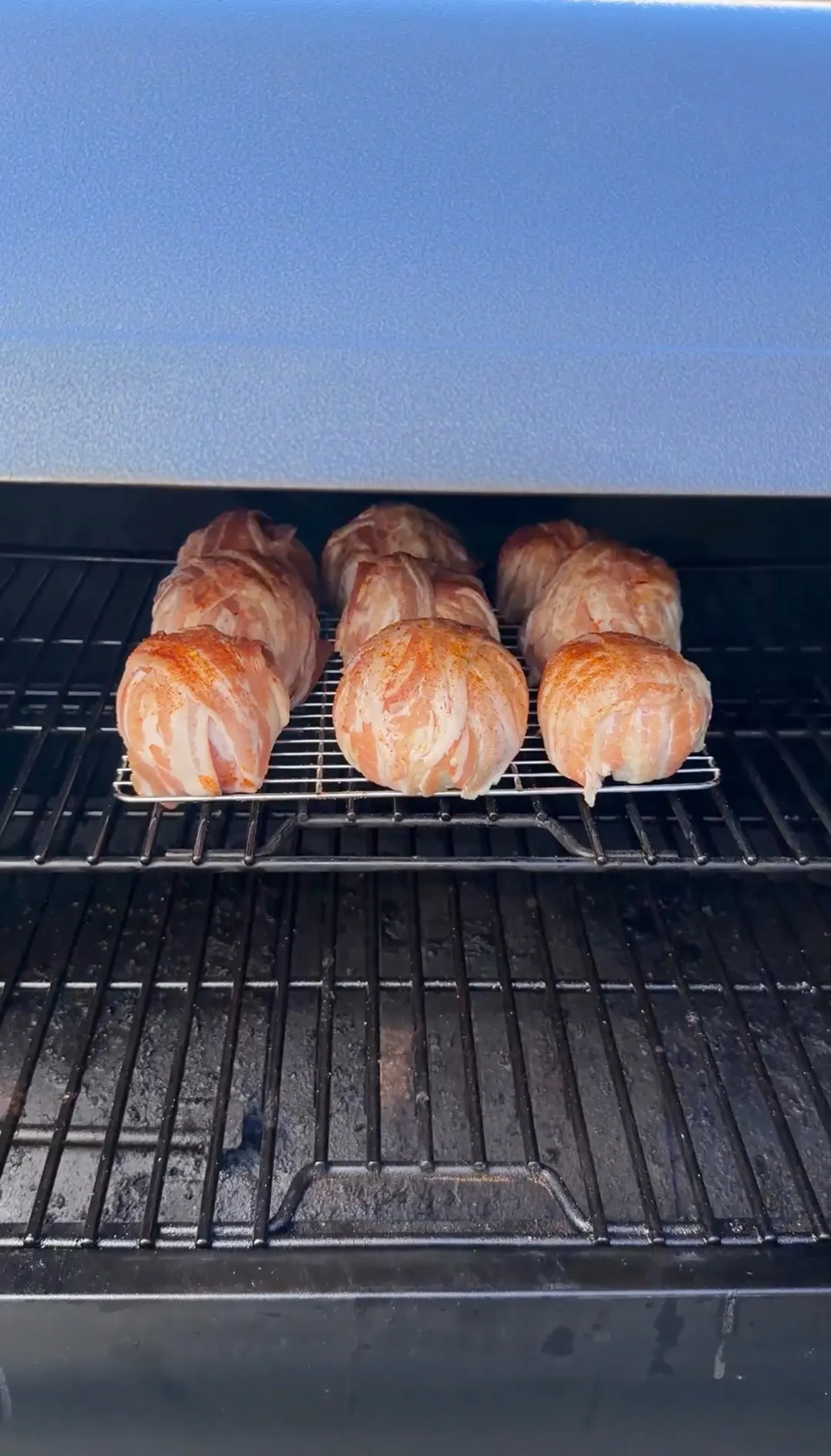 Six bacon-wrapped onion bombs seasoned with spices cooking on a wire rack inside a pellet smoker