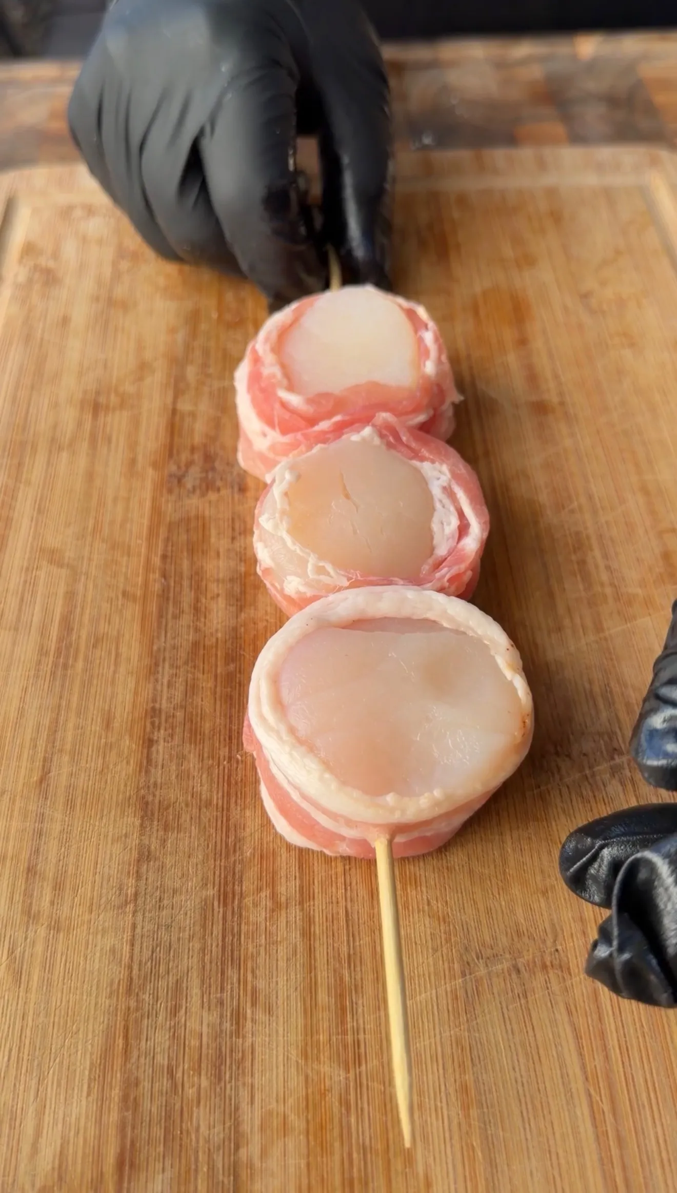 Three raw bacon-wrapped scallops threaded on a wooden skewer on a cutting board, prepared by gloved hands