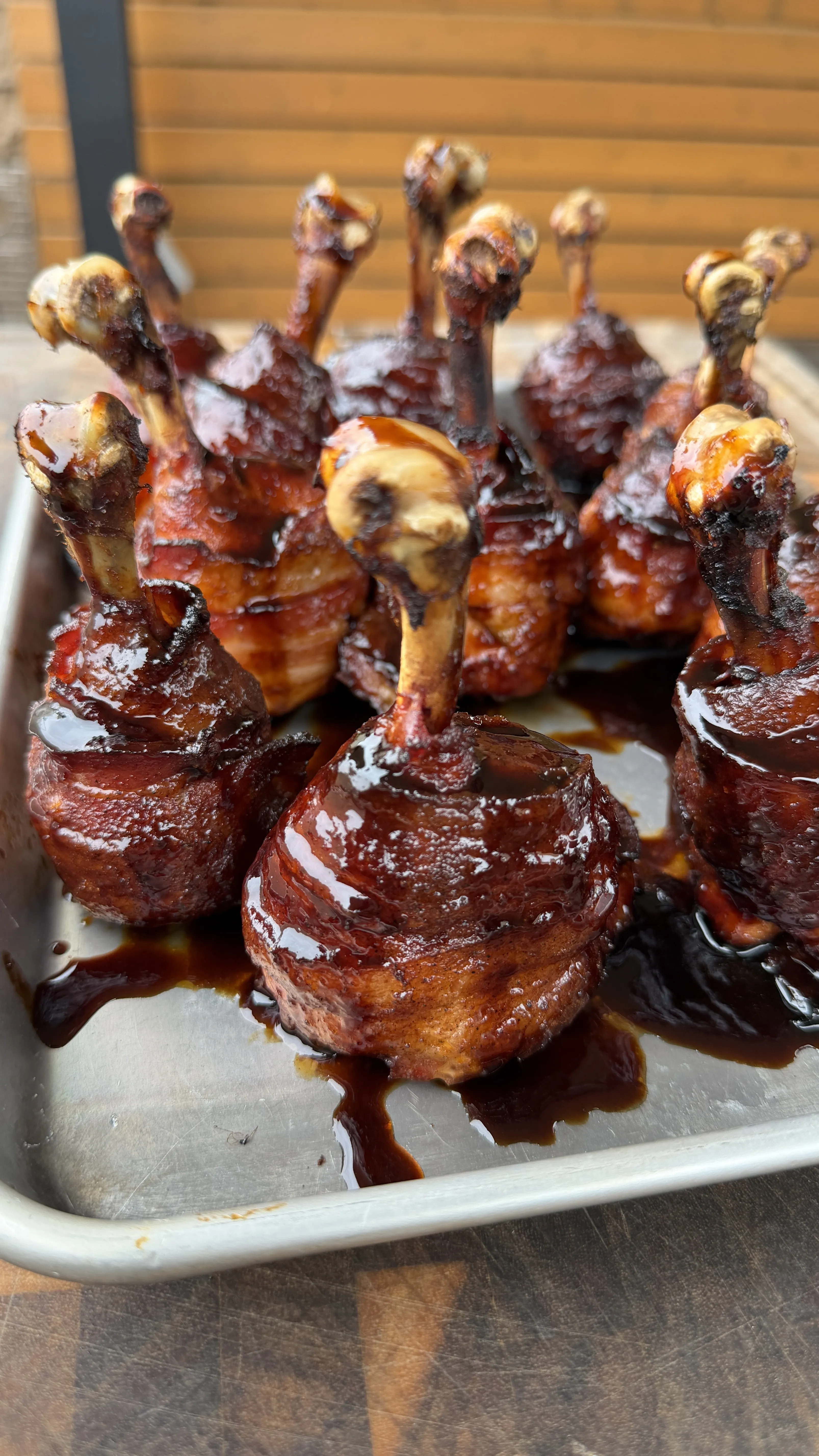 Bacon wrapped hot and spicy chicken lollipops coated in glossy BBQ sauce on a metal tray