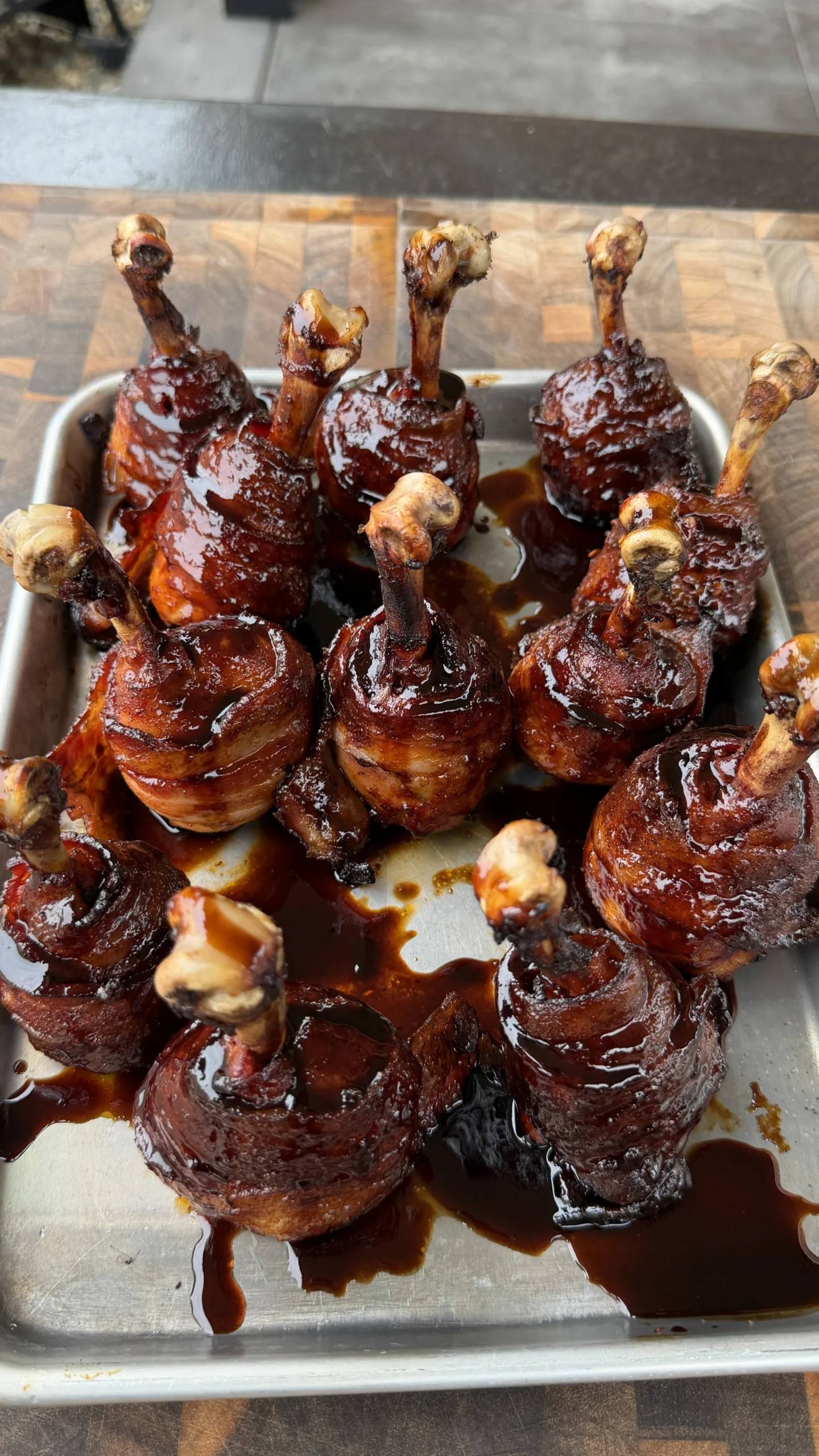 Bacon wrapped chicken lollipops coated in glossy BBQ sauce on a stainless steel tray