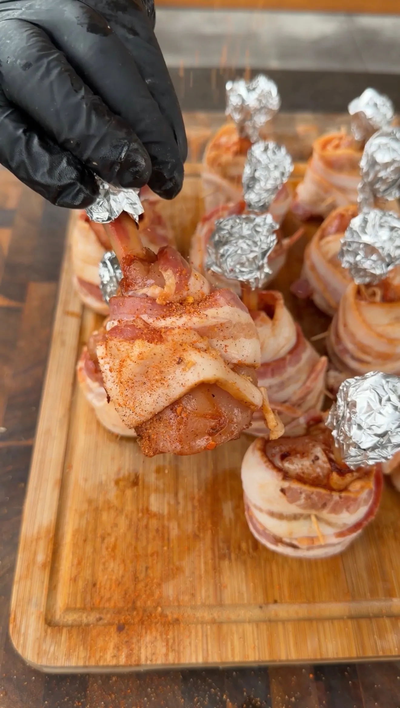 Bacon-wrapped spicy chicken lollipops with foil-tipped bones on a wooden cutting board, seasoned with red spice rub