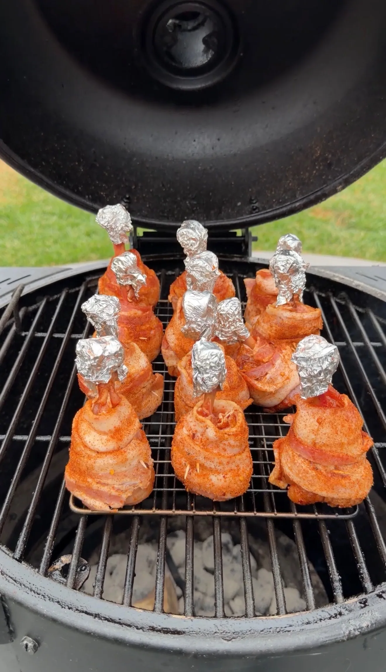 Bacon-wrapped spicy chicken lollipops with foil-covered bones smoking on a charcoal grill