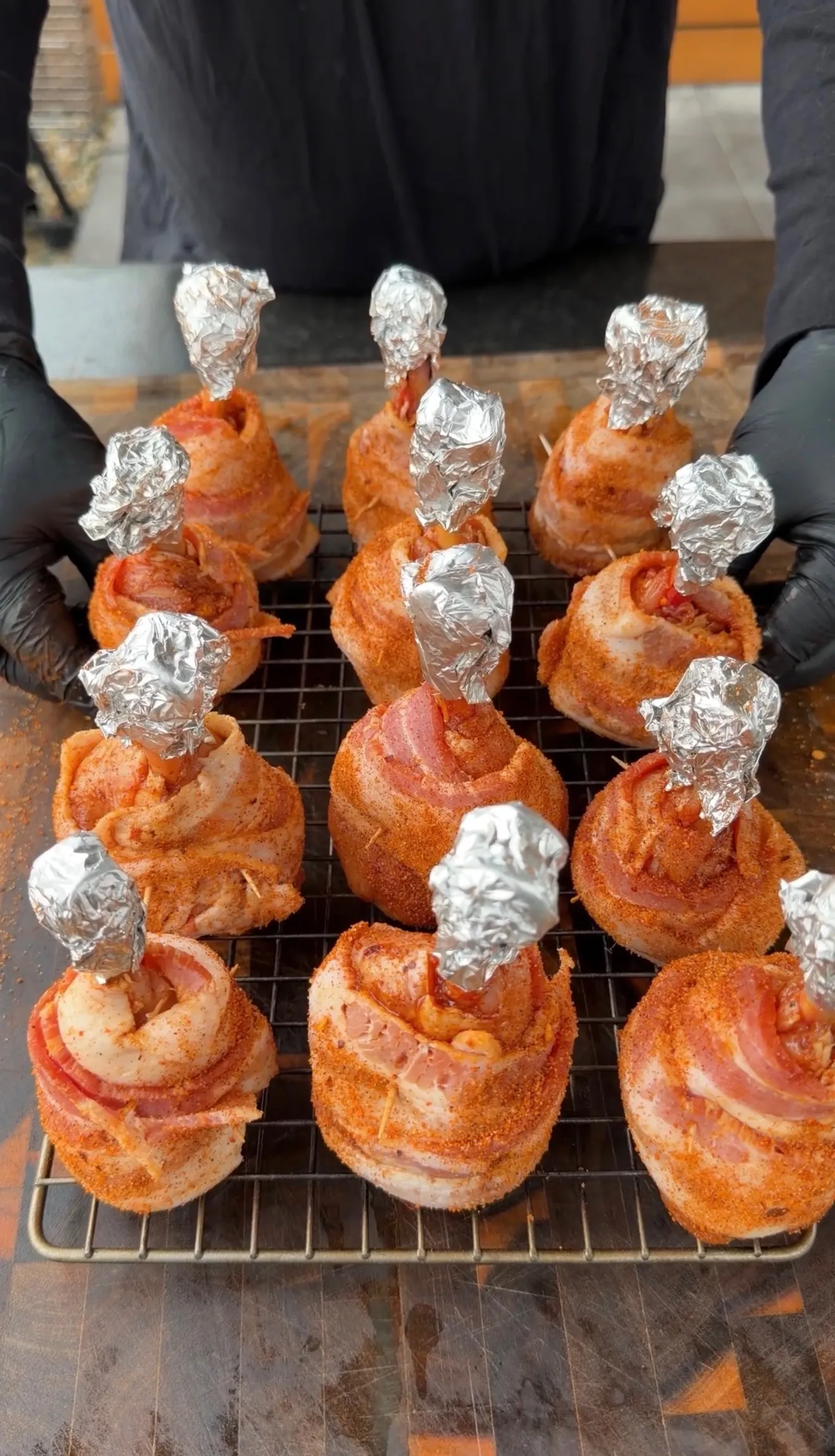 12 bacon-wrapped spicy chicken lollipops coated in red seasoning with foil-covered bones on a wire rack