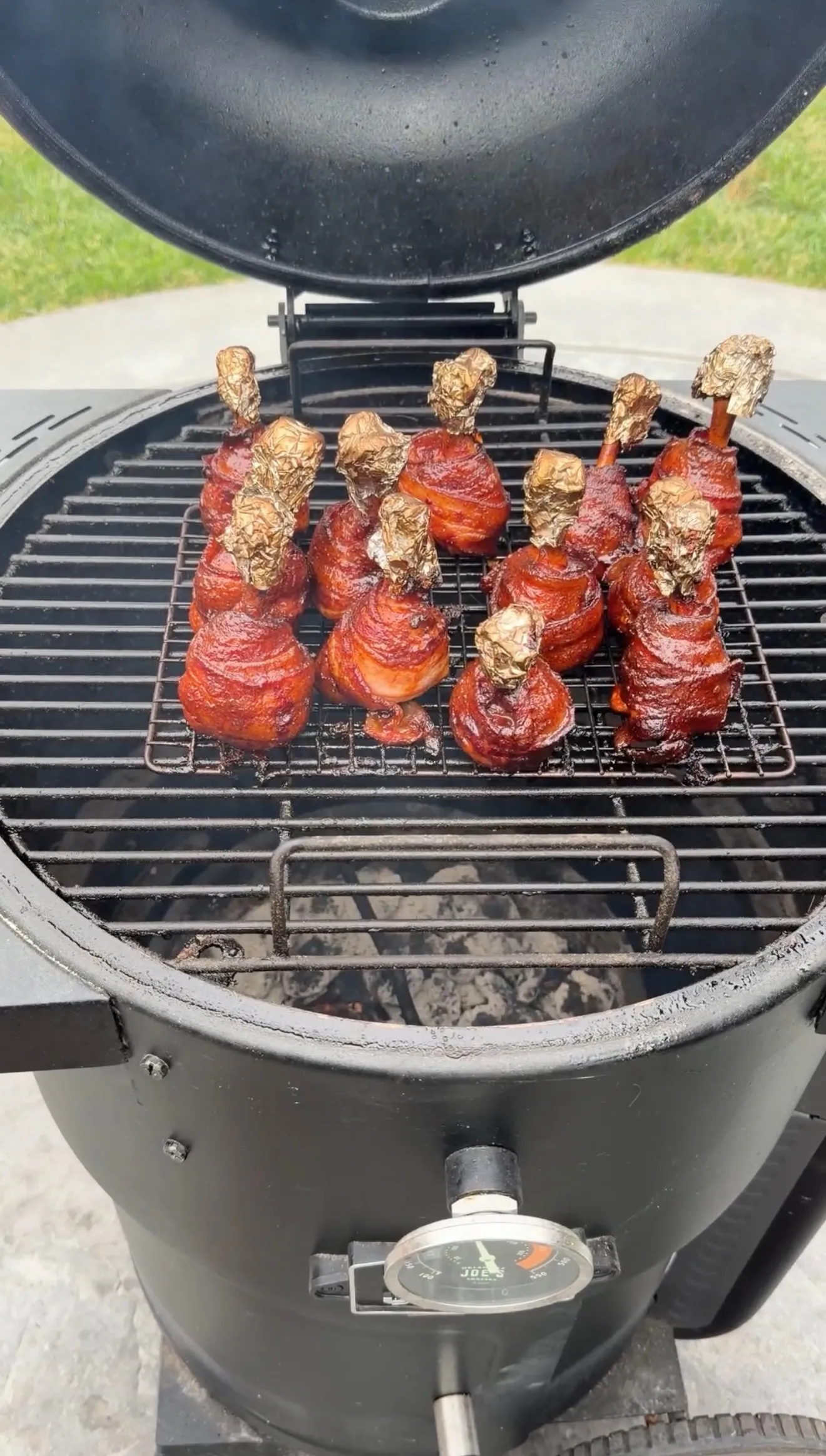 Bacon-wrapped hot and spicy chicken lollipops with foil-tipped bones smoking on a Kamado Joe charcoal grill