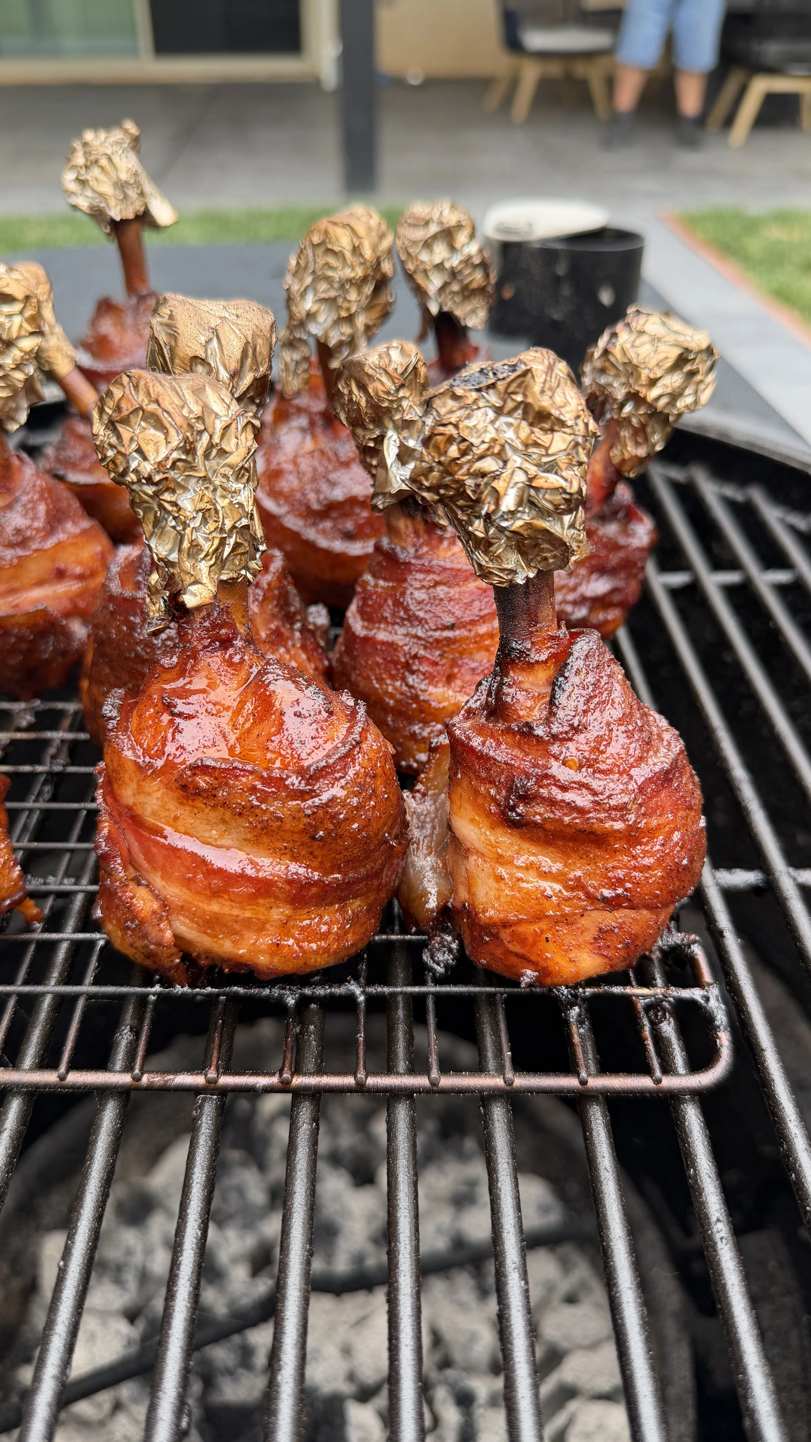 Bacon-wrapped hot and spicy chicken lollipops with foil-tipped bones smoking on a charcoal grill