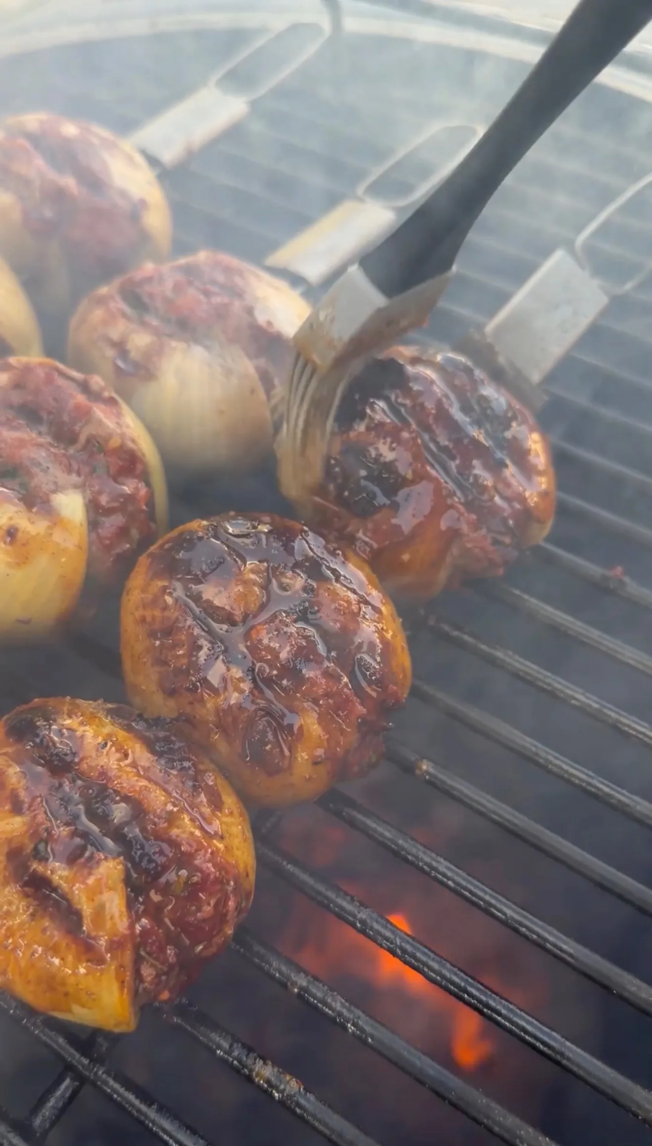 BBQ-glazed stuffed onion bombs sizzling on a charcoal grill with flames, being basted with sauce