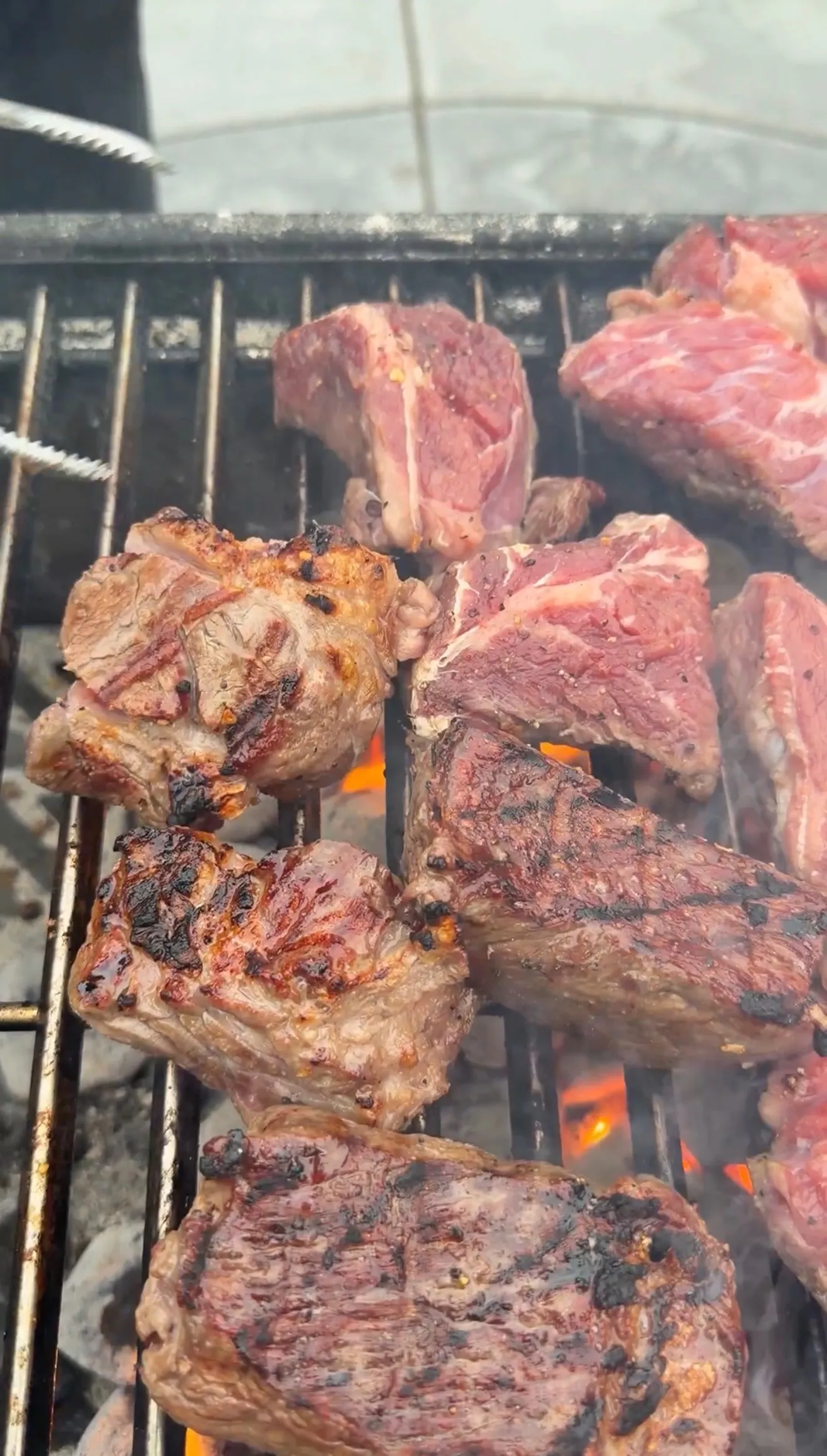 Seasoned birria beef chunks at various stages of grilling over open flames on a charcoal grill