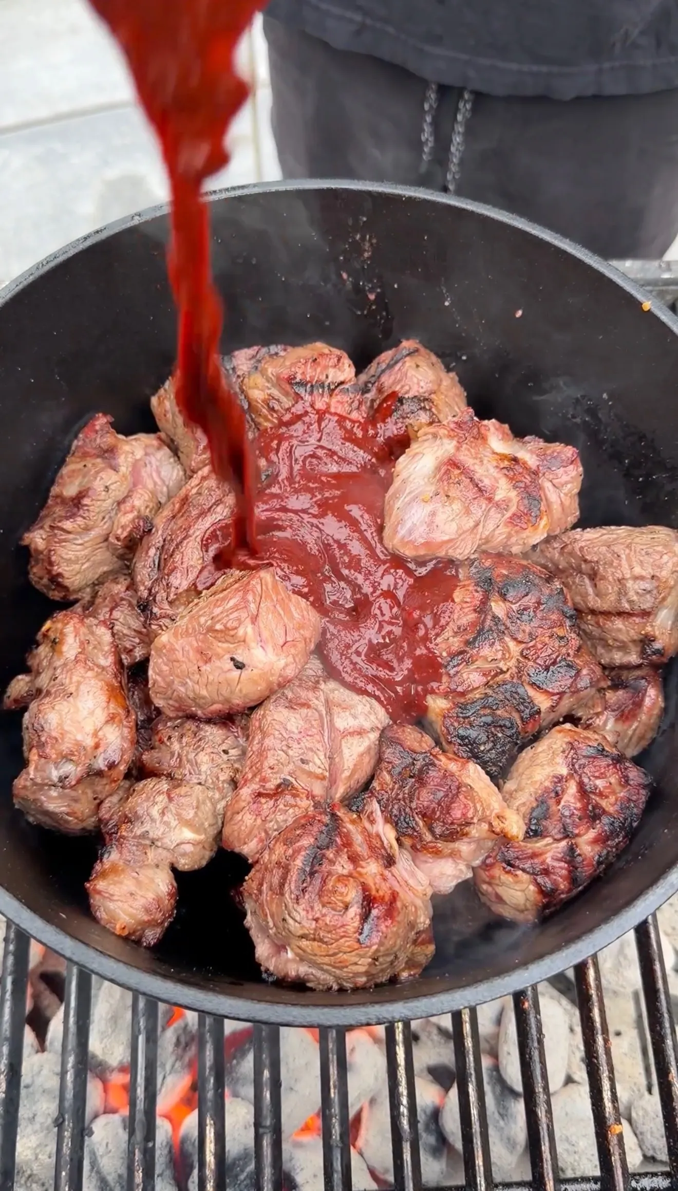 Seared birria beef chunks in cast iron pan over charcoal grill with red guajillo sauce being poured over