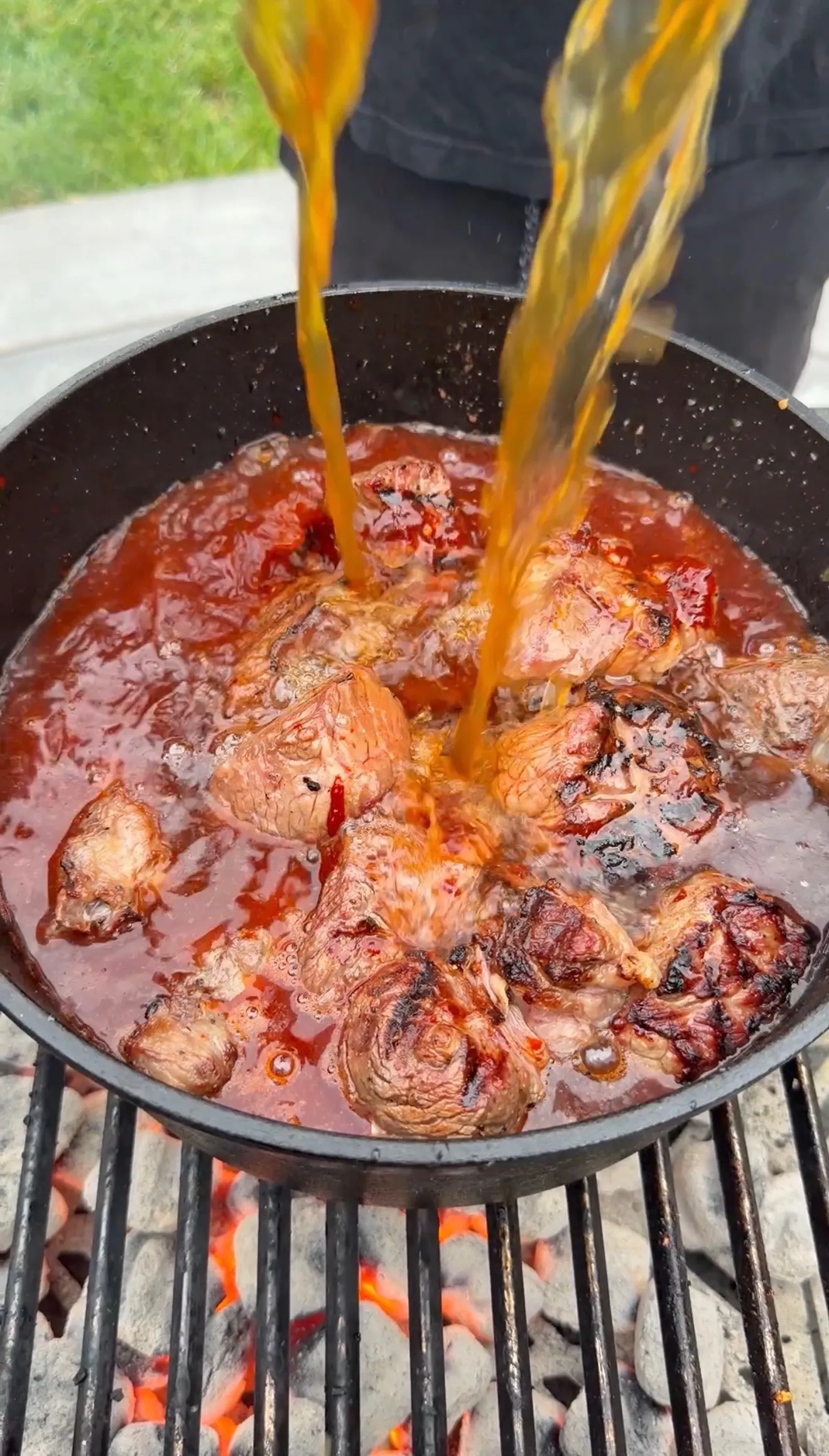 Chunks of seared birria beef simmering in rich red consommé broth in a cast iron pan over charcoal fire