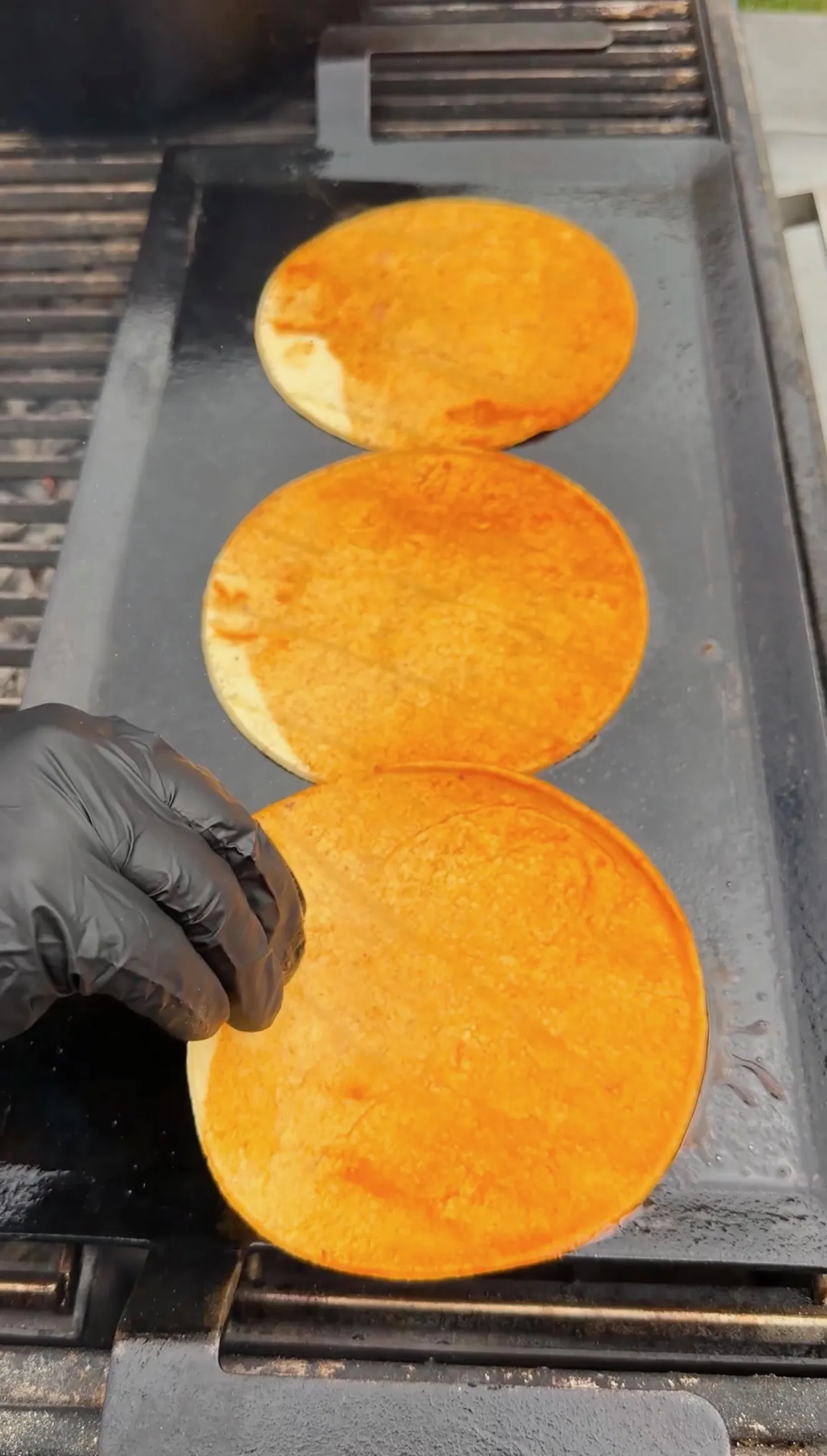 Three orange-tinted birria taco tortillas toasting on a flat iron griddle, held by a gloved hand