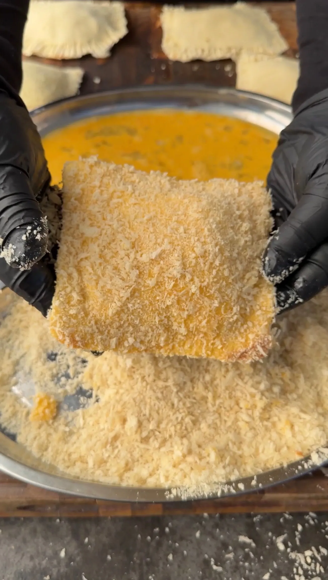 Hands in black gloves coating a cheesesteak stuffed pocket in panko breadcrumbs over egg wash bowl