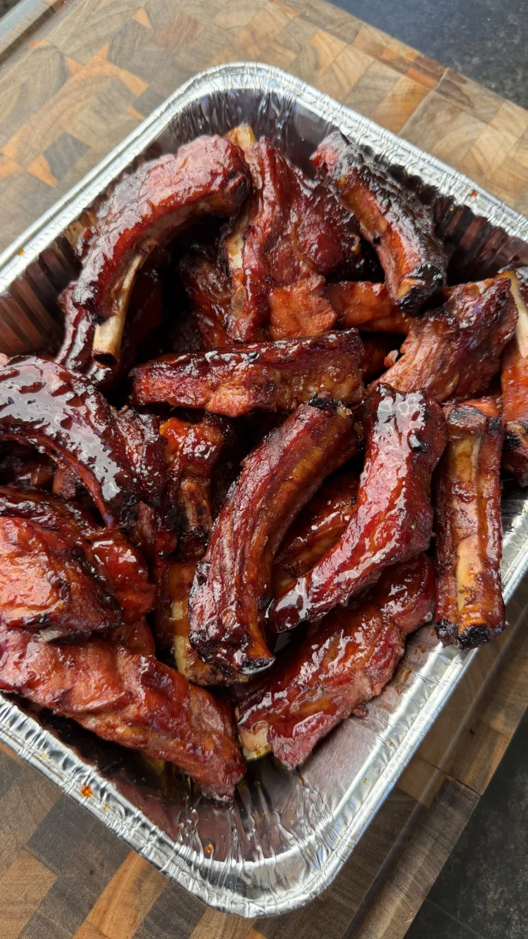 Glazed brown sugar bourbon party ribs piled in a foil pan on a wooden cutting board