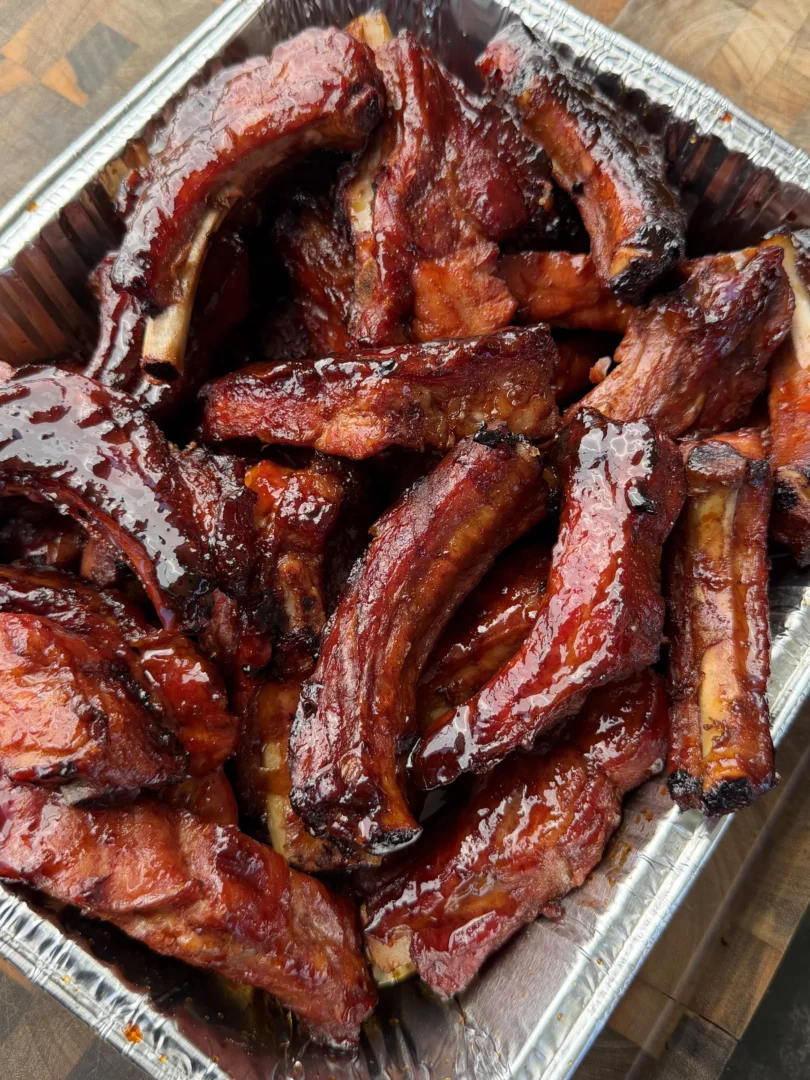 Glazed brown sugar bourbon party ribs piled in a foil pan on a wooden cutting board