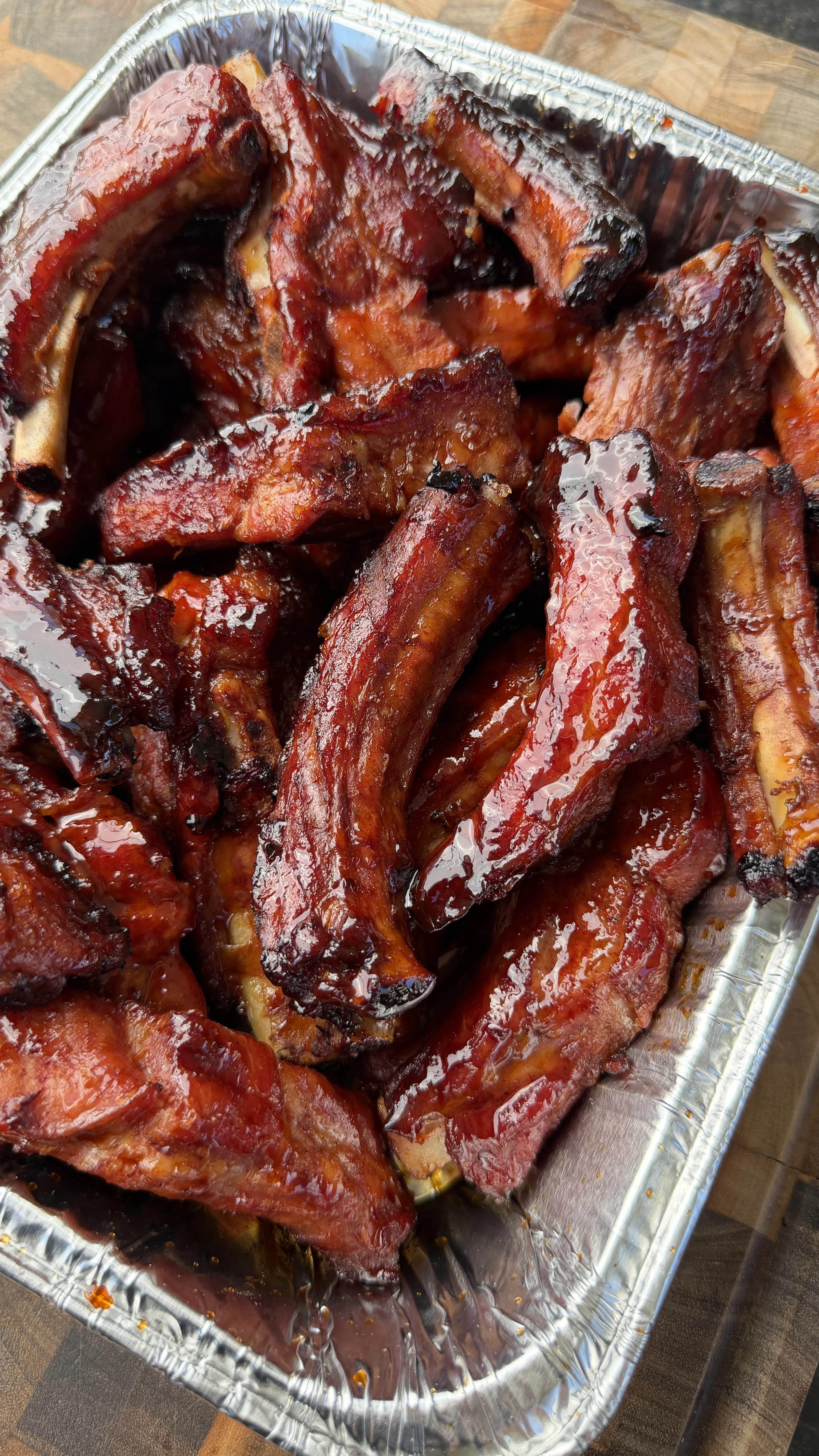 Glossy brown sugar bourbon party ribs piled in a foil pan with caramelized BBQ glaze