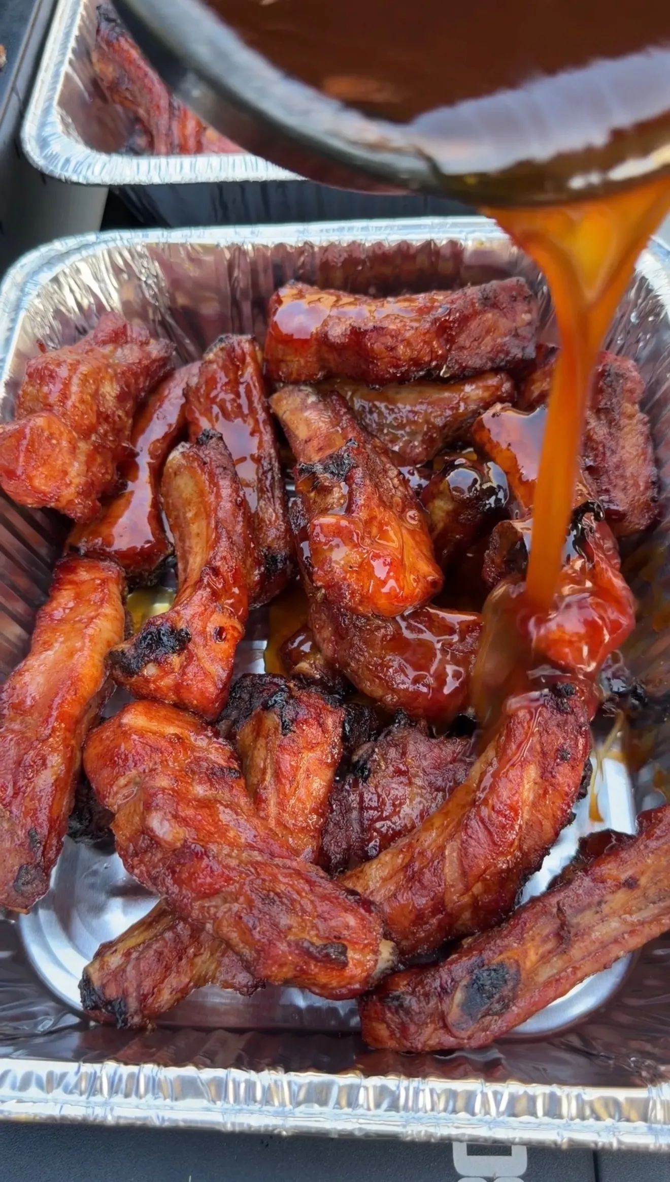 BBQ brown sugar bourbon party ribs piled in a foil pan with glossy sauce being poured over top