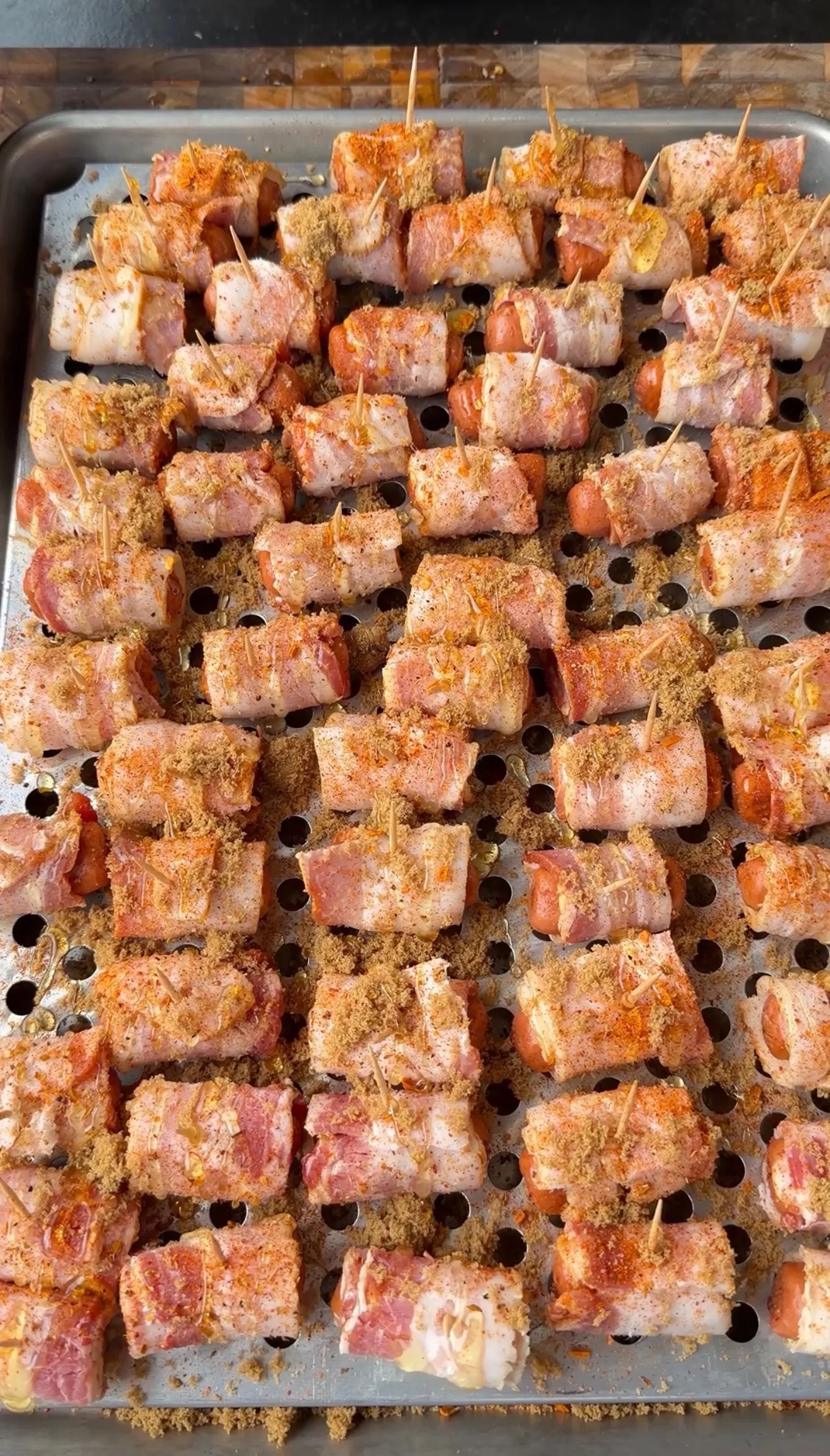 Raw bacon-wrapped little smokies seasoned with Cajun spices and brown sugar on a perforated baking sheet