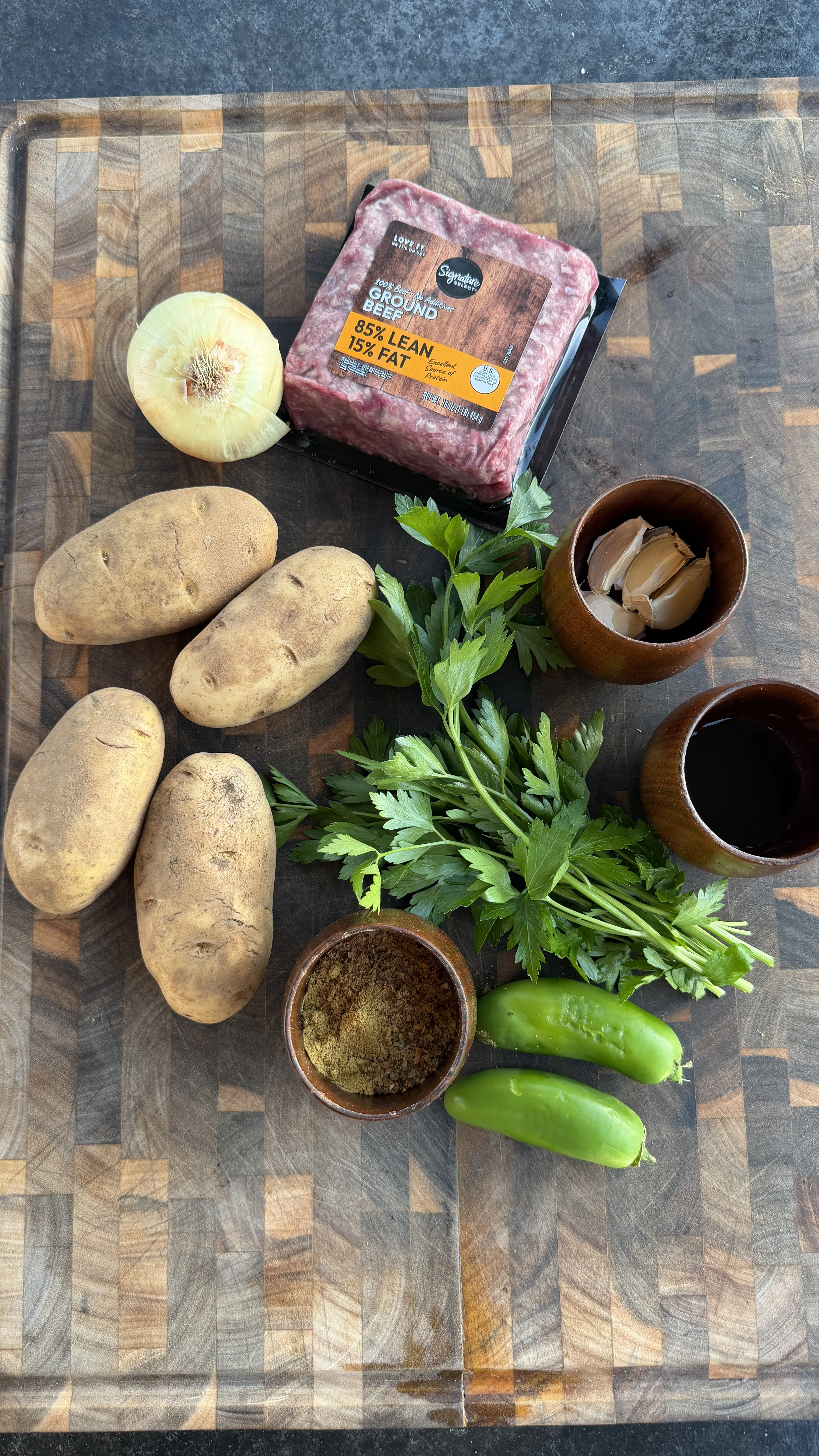 Raw ingredients for Cajun butter potato beef kebabs including ground beef, russet potatoes, onion, jalapeños, parsley, garlic, and spices on a wooden board
