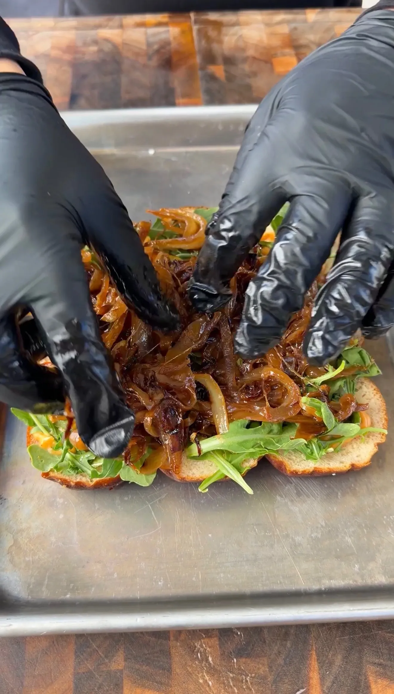 Gloved hands layering caramelized onions and arugula onto tri-tip cowboy butter sliders in a metal pan
