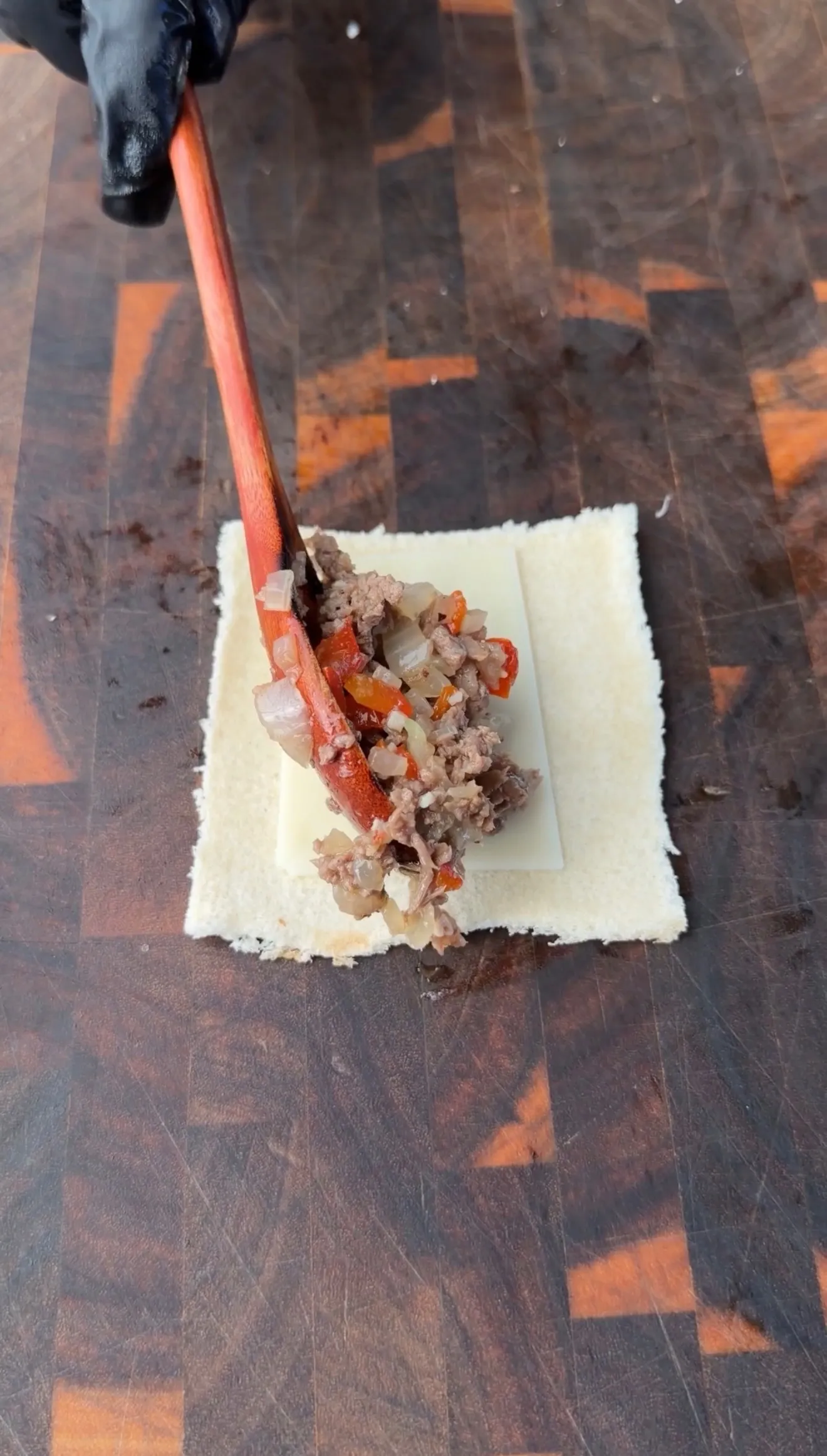 Meat and vegetable cheesesteak filling being spooned onto a square of pastry dough on a wooden cutting board