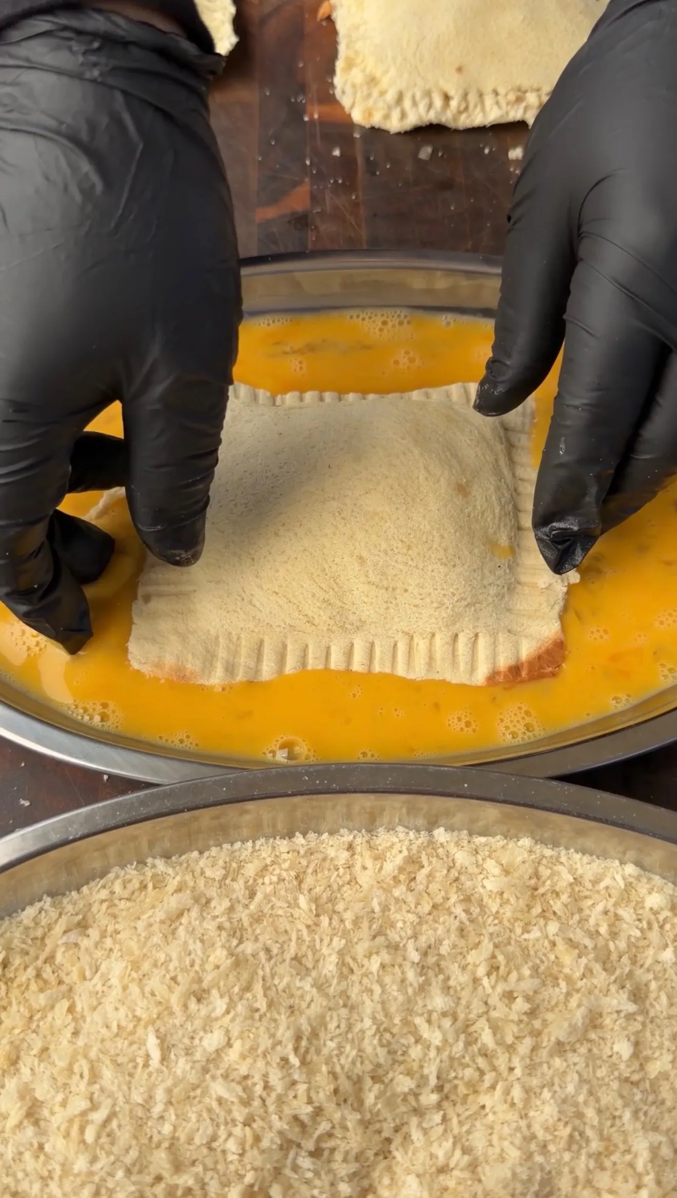 Hands in black gloves dipping a stuffed cheesesteak pocket into egg wash before coating in panko breadcrumbs